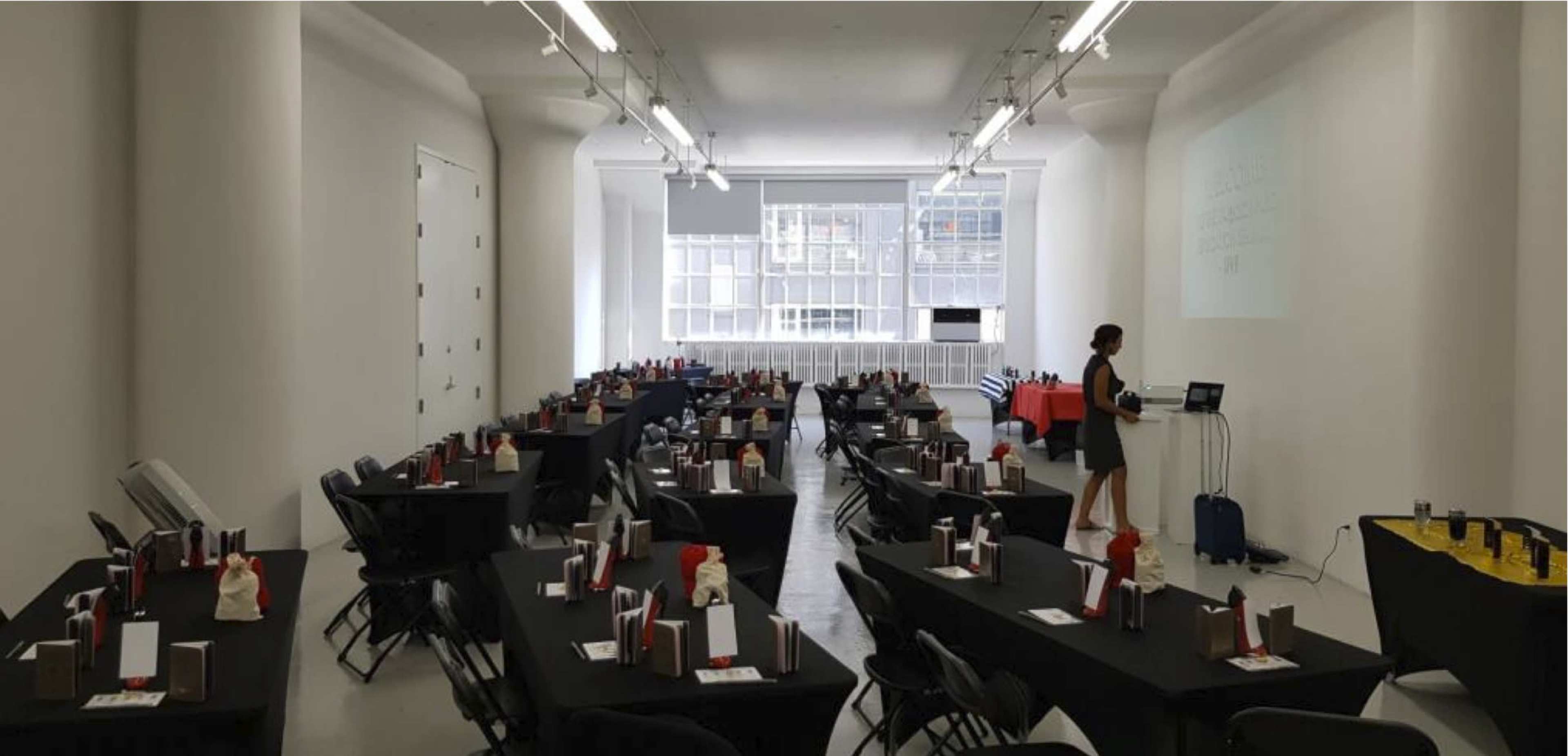 A spacious event room is set up with several tables covered in black cloth and arranged with red and white decorations, while a presenter stands near a projector screen at the front.