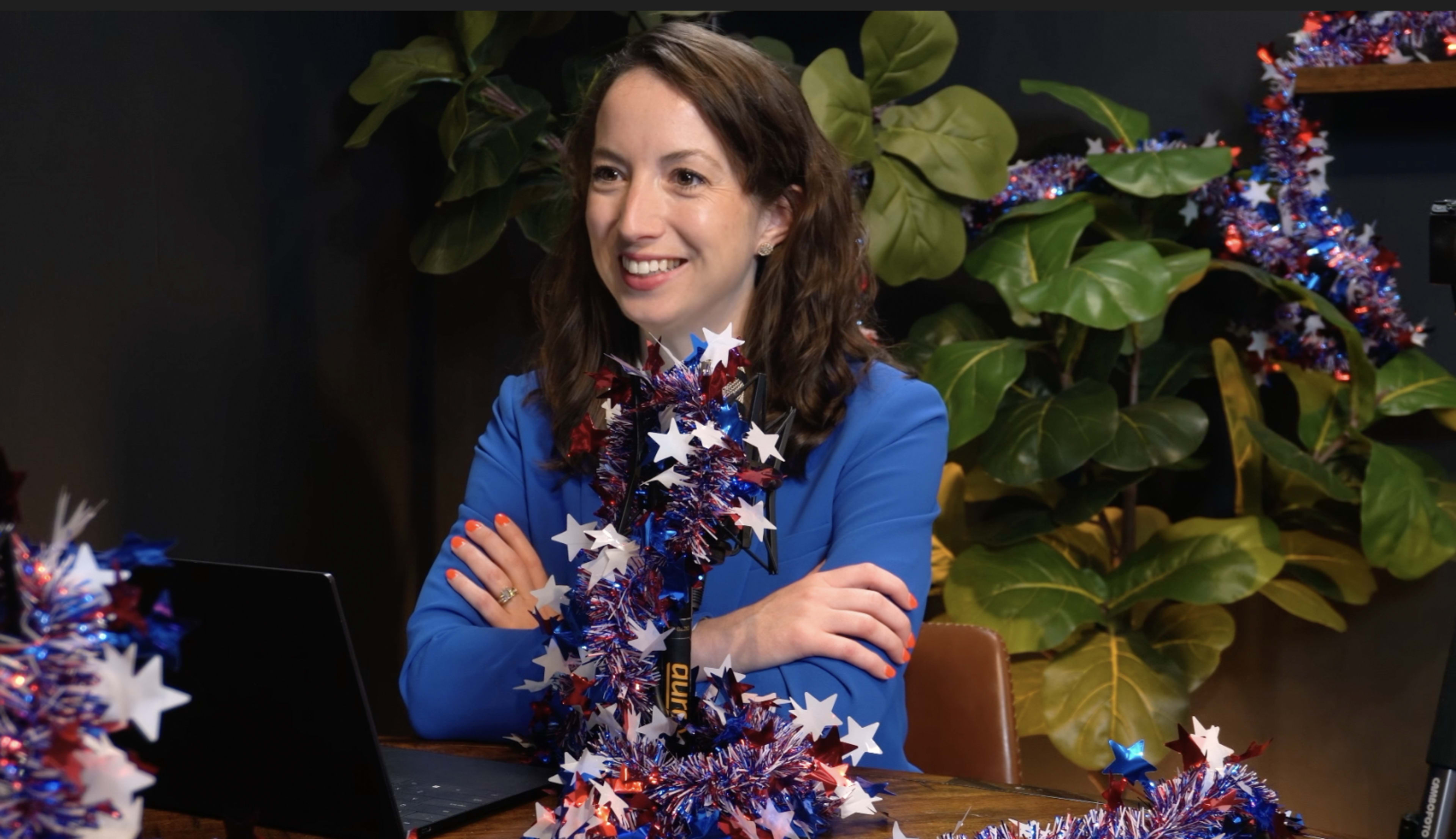 A woman with brown hair seated at a table adorned with festive decorations and plants, smiling while looking at a laptop.
