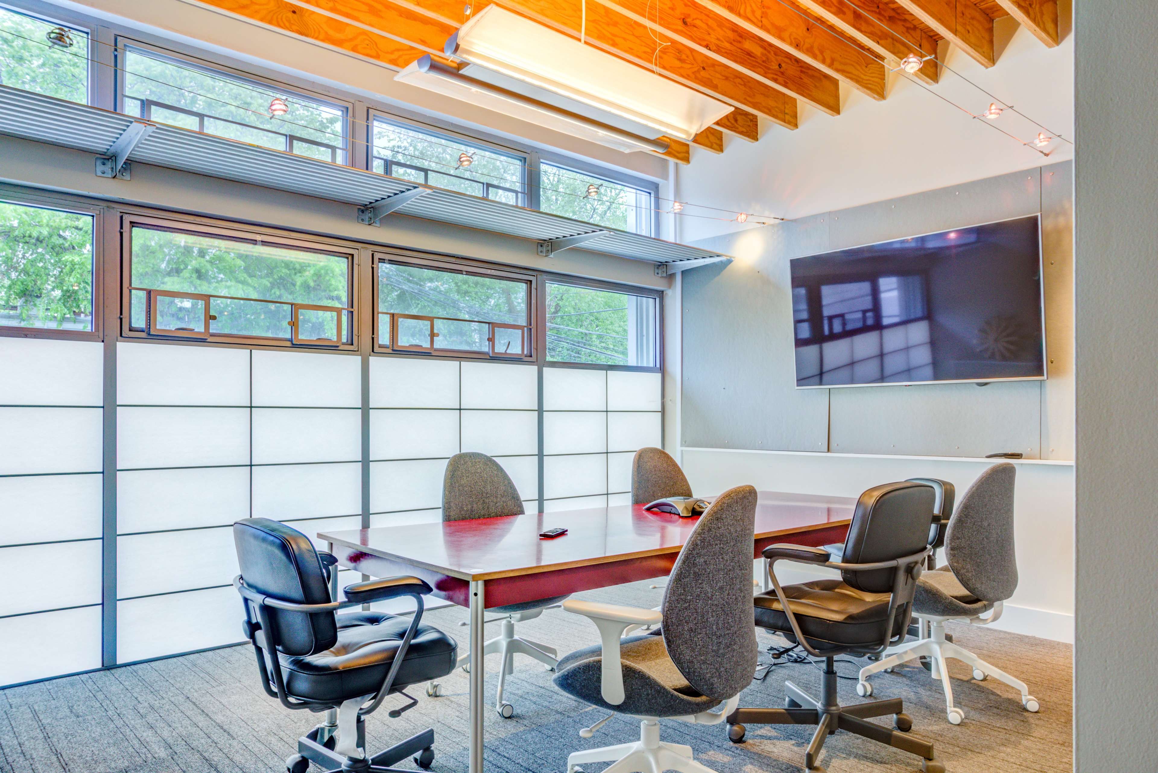 A modern conference room features a large table surrounded by rolling chairs, large windows, and a screen mounted on the wall.