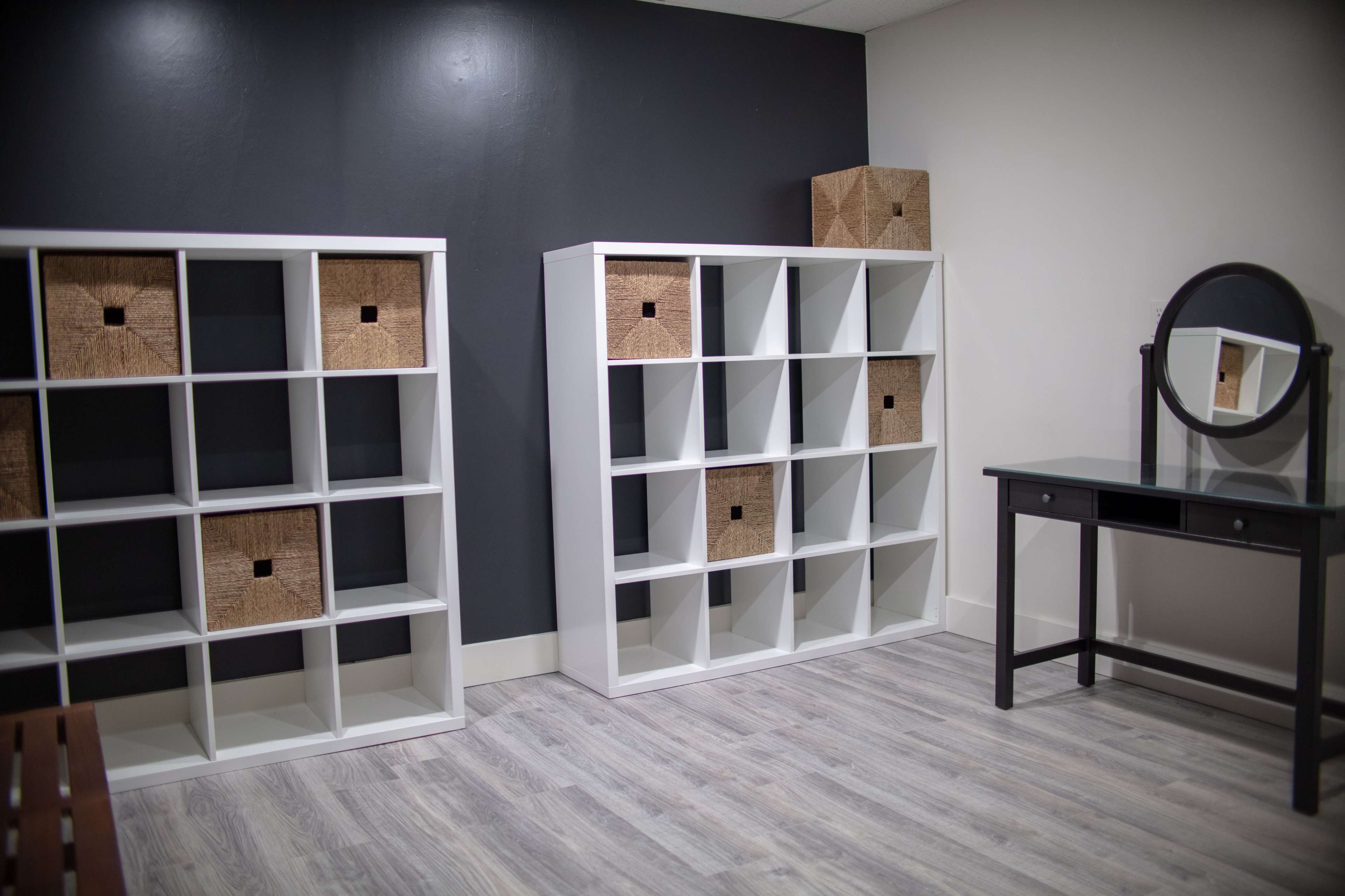 A simple room with two white shelving units, each containing woven storage boxes, and a black vanity table with a round mirror.