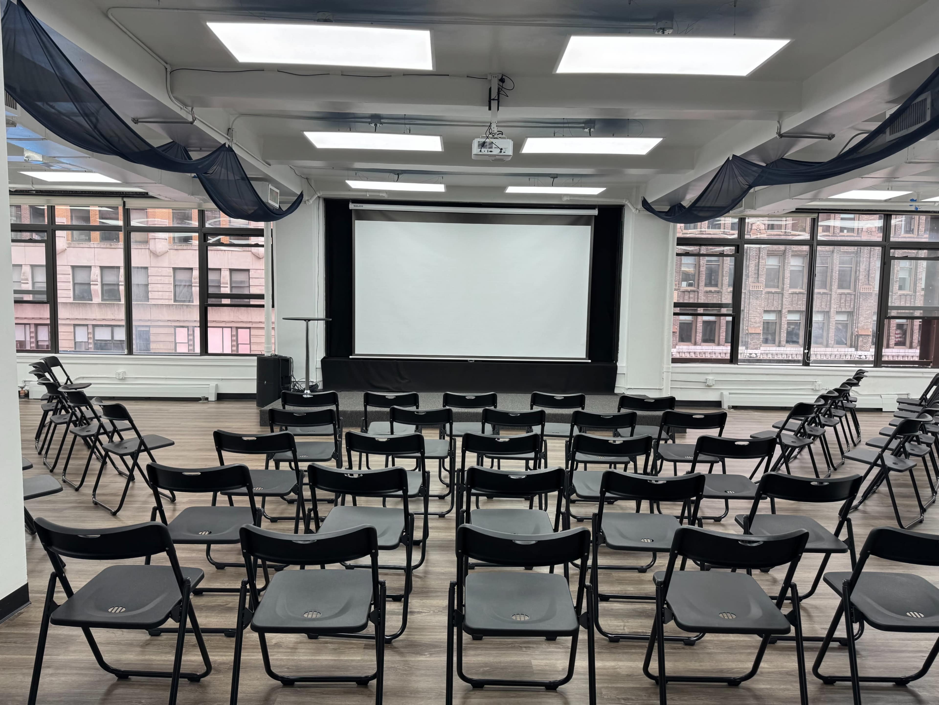 A large room is set up with rows of black folding chairs facing a blank projector screen.
