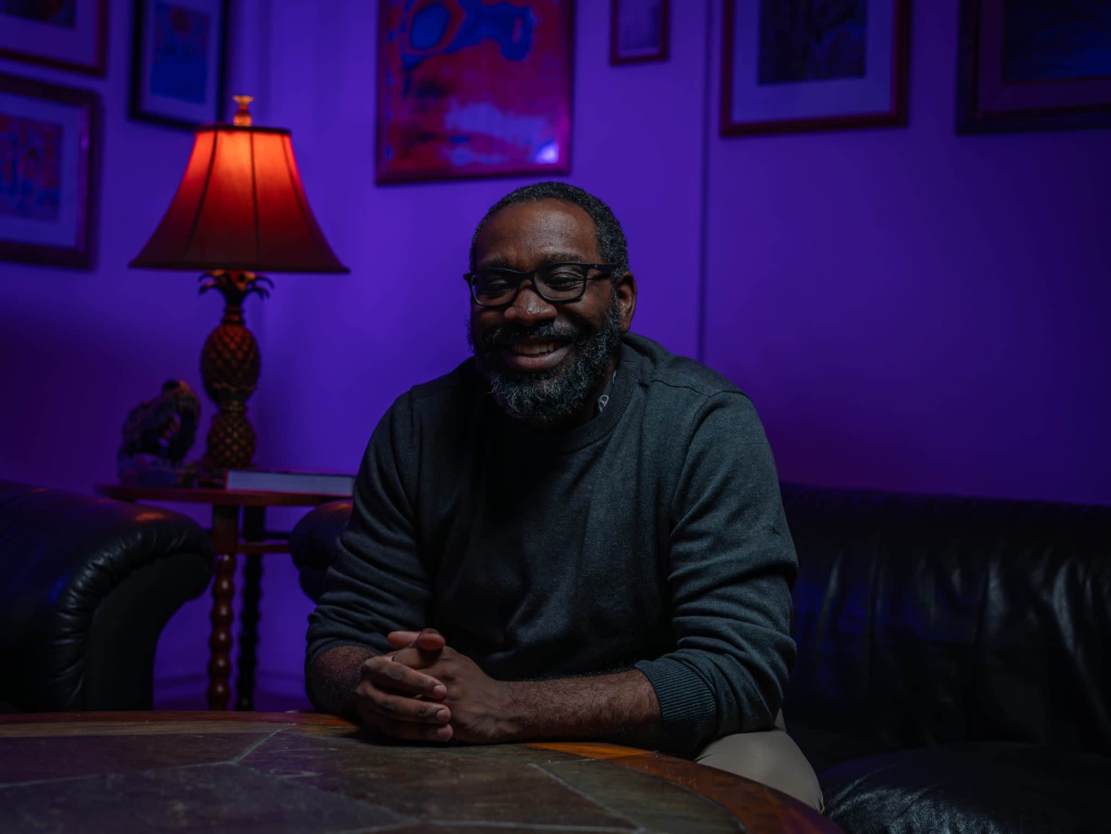 A man with a beard sits on a leather couch, smiling in a dimly lit room with purple and warm orange lighting.