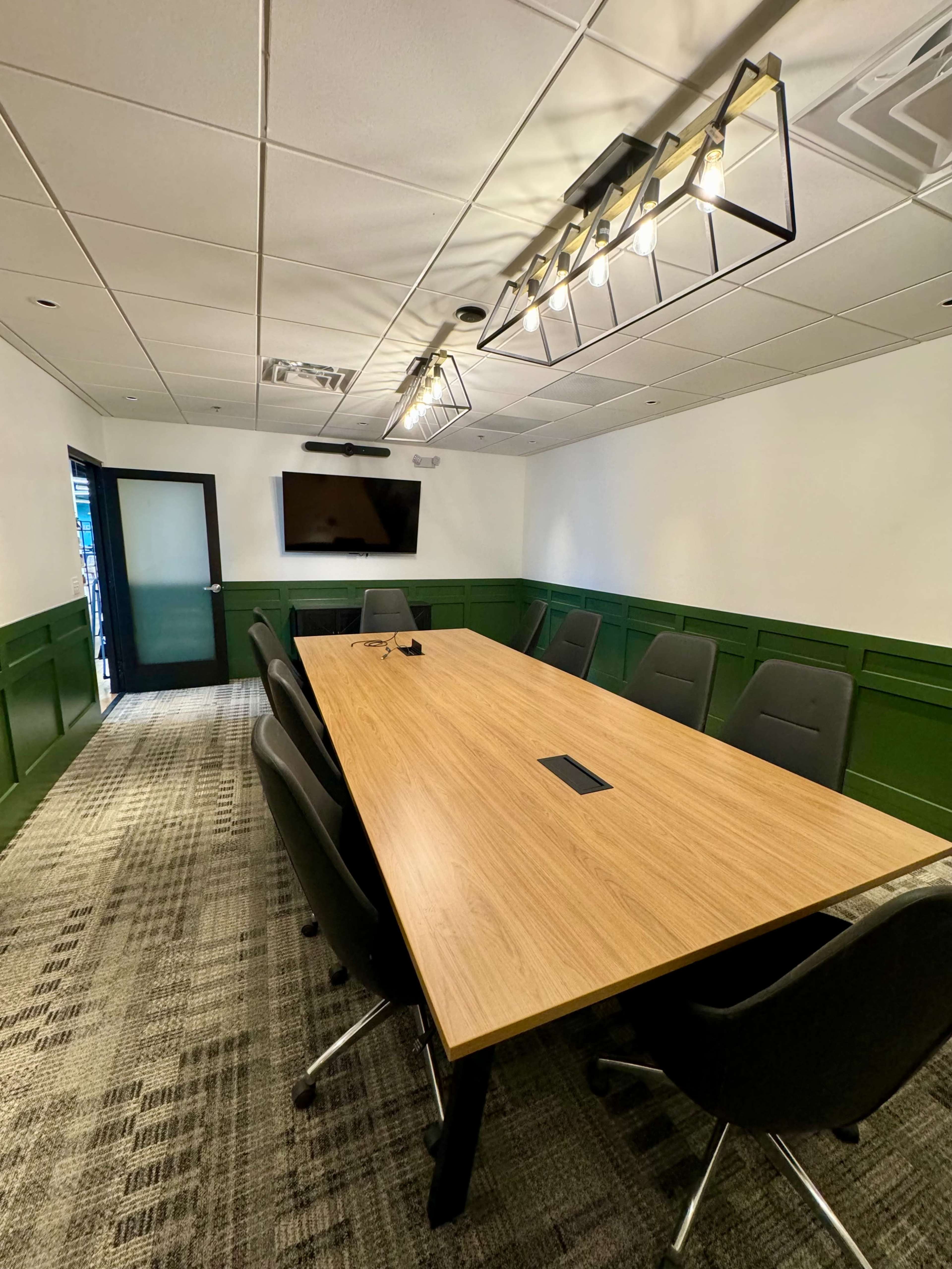 The image shows a modern conference room with a long wooden table, black chairs, and a wall-mounted television.