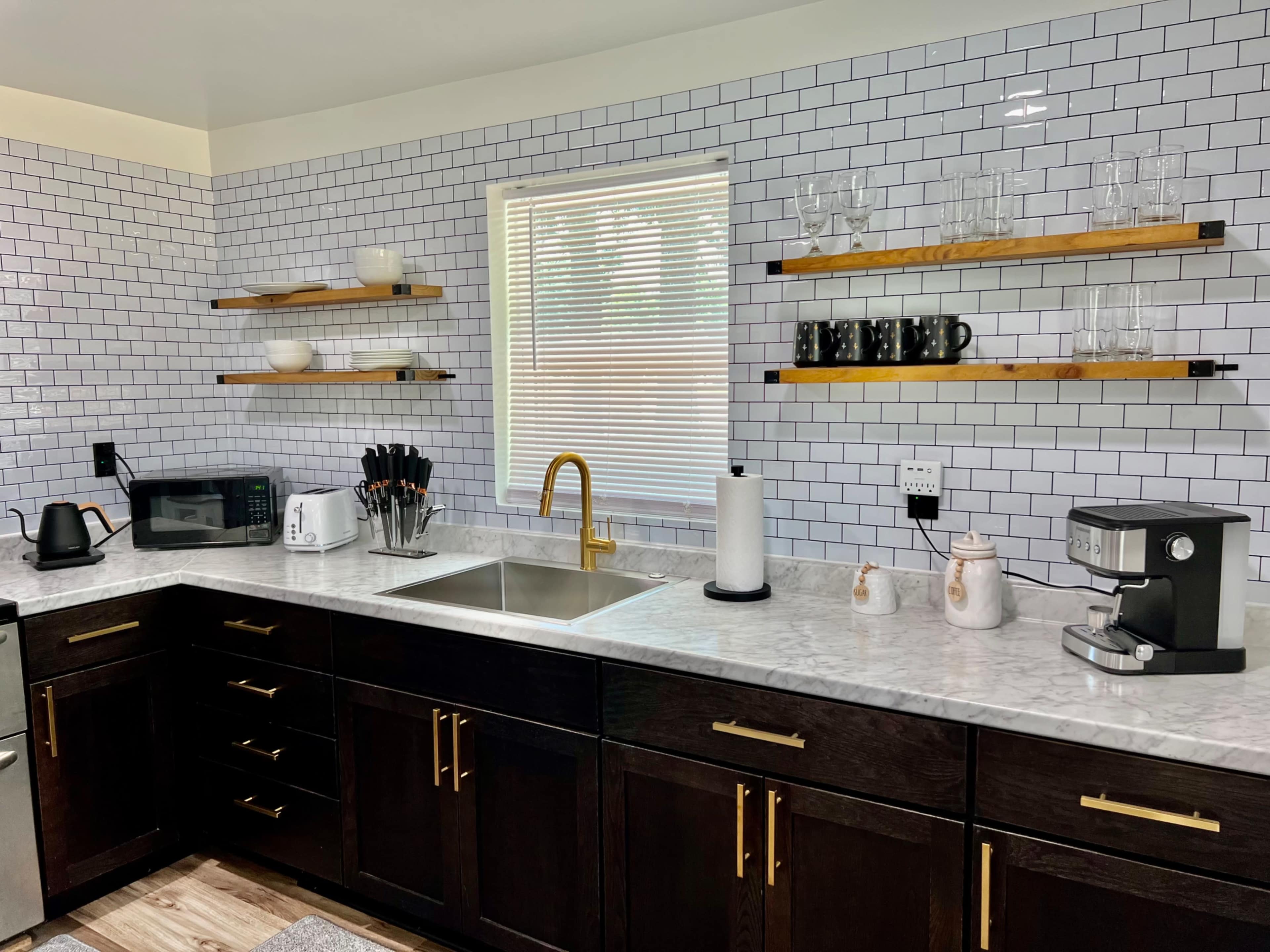 A modern kitchen with dark cabinets, a marble countertop, open shelving, and various appliances including a microwave, kettle, and coffee machine.