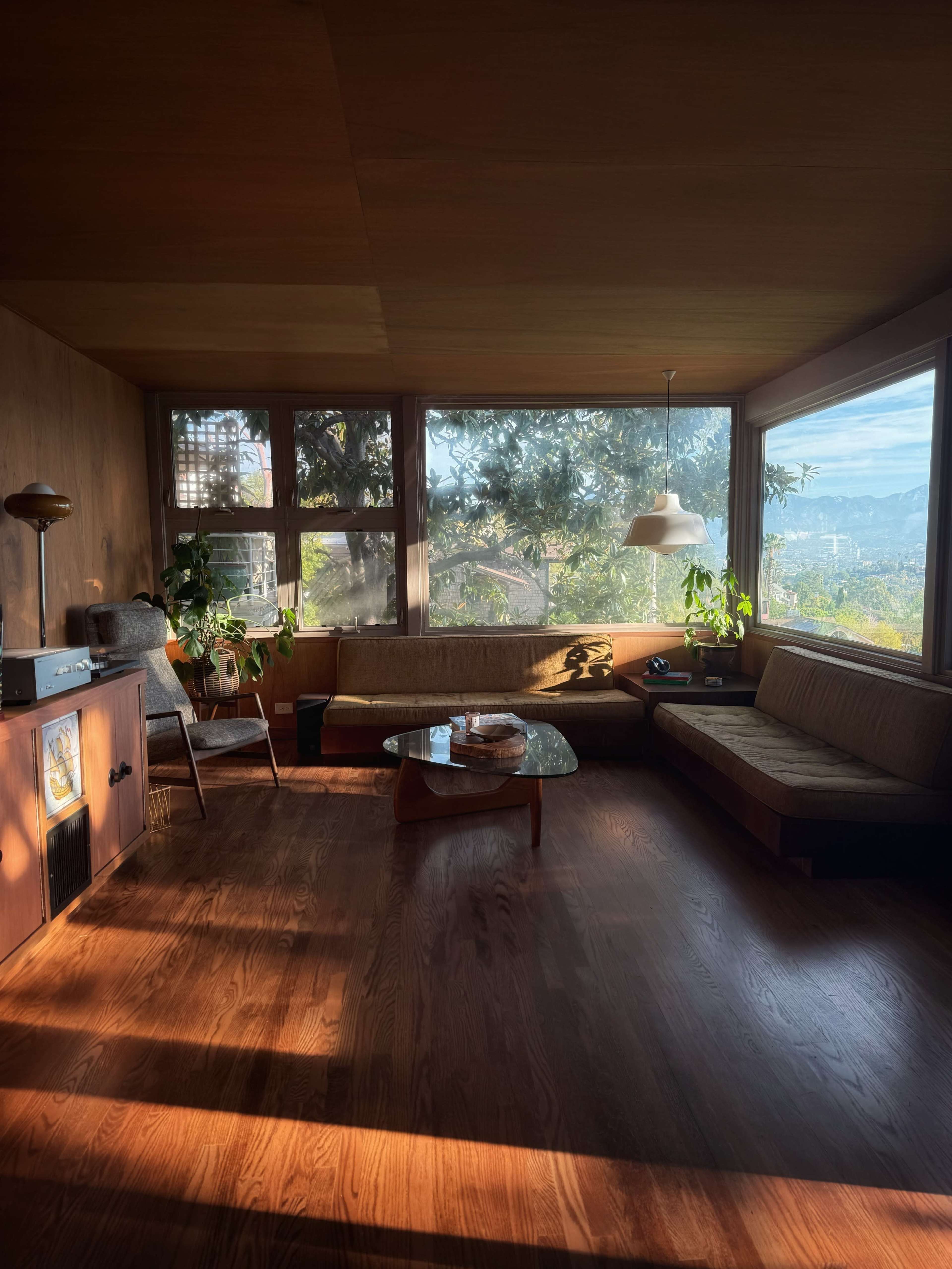 A sunlit, sparsely furnished living room features large windows, wooden walls, a glass coffee table, and a corner chair.