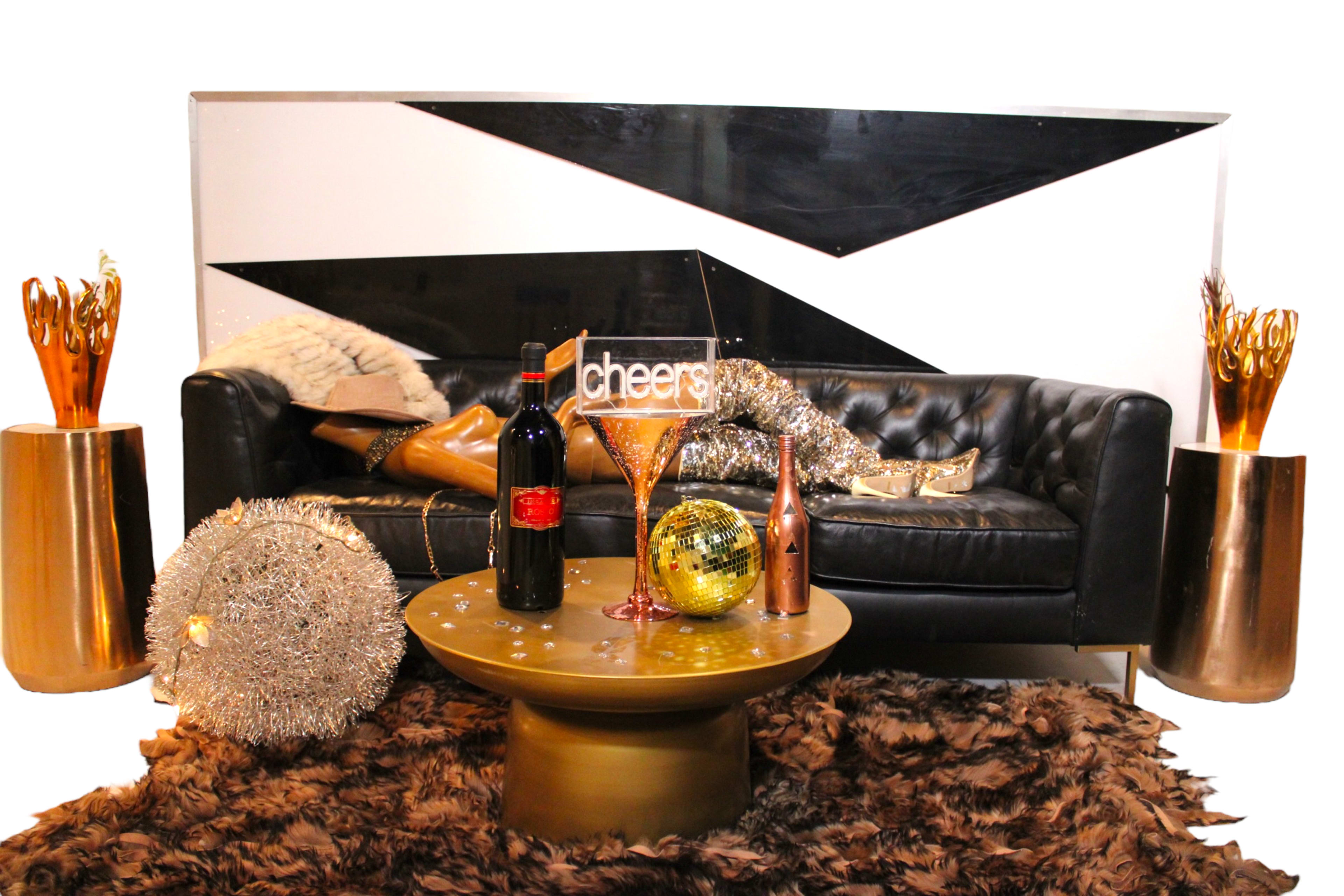 A black leather sofa is placed against a decorative wall, with a gold table in front featuring wine bottles, a glass, and decorative items like a disco ball and a sign that reads "cheers."