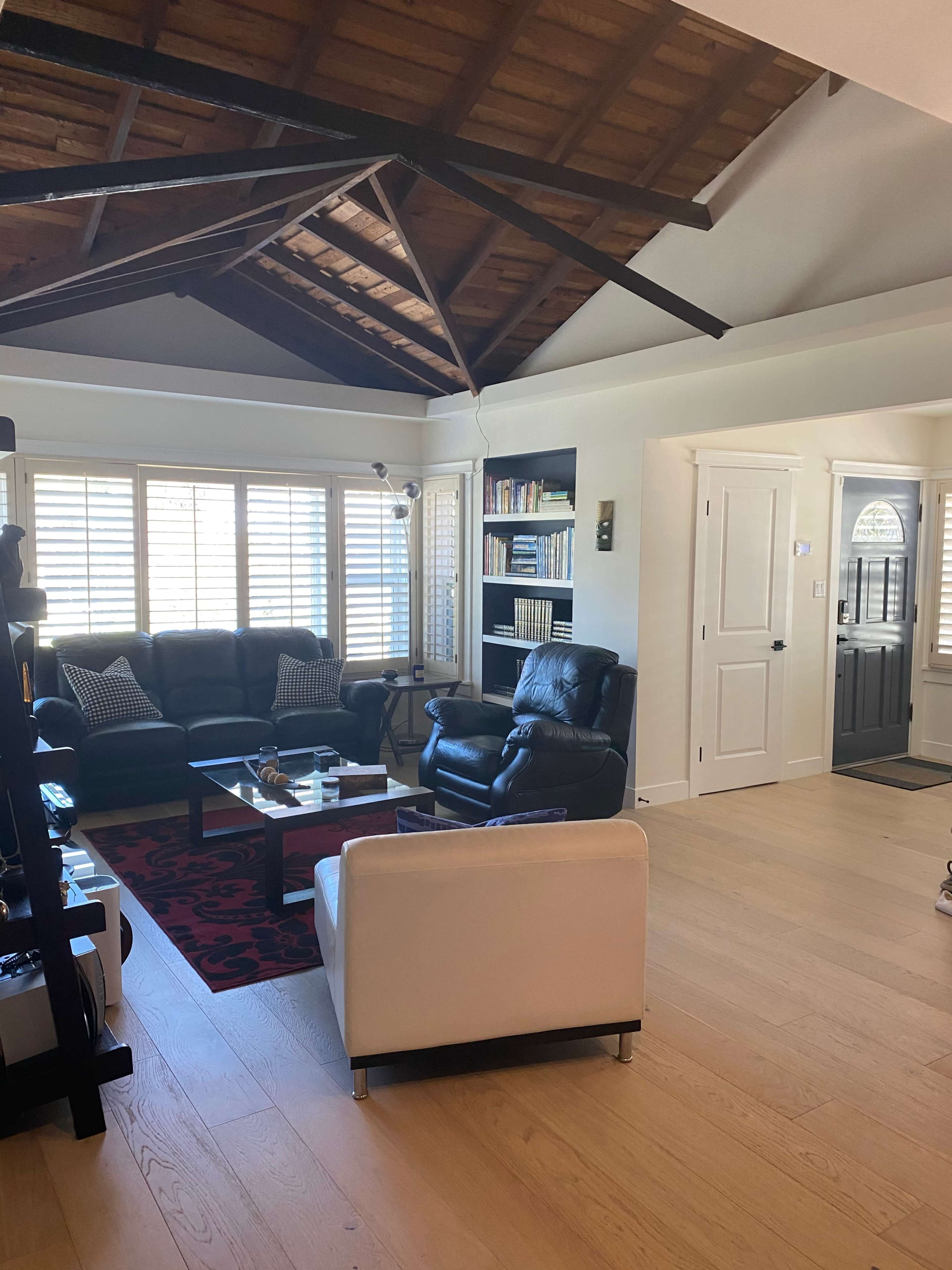The image shows a living room with a vaulted ceiling, furnished with a black leather sofa, an armchair, a white club chair, and a coffee table, while windows with shutters let in natural light.