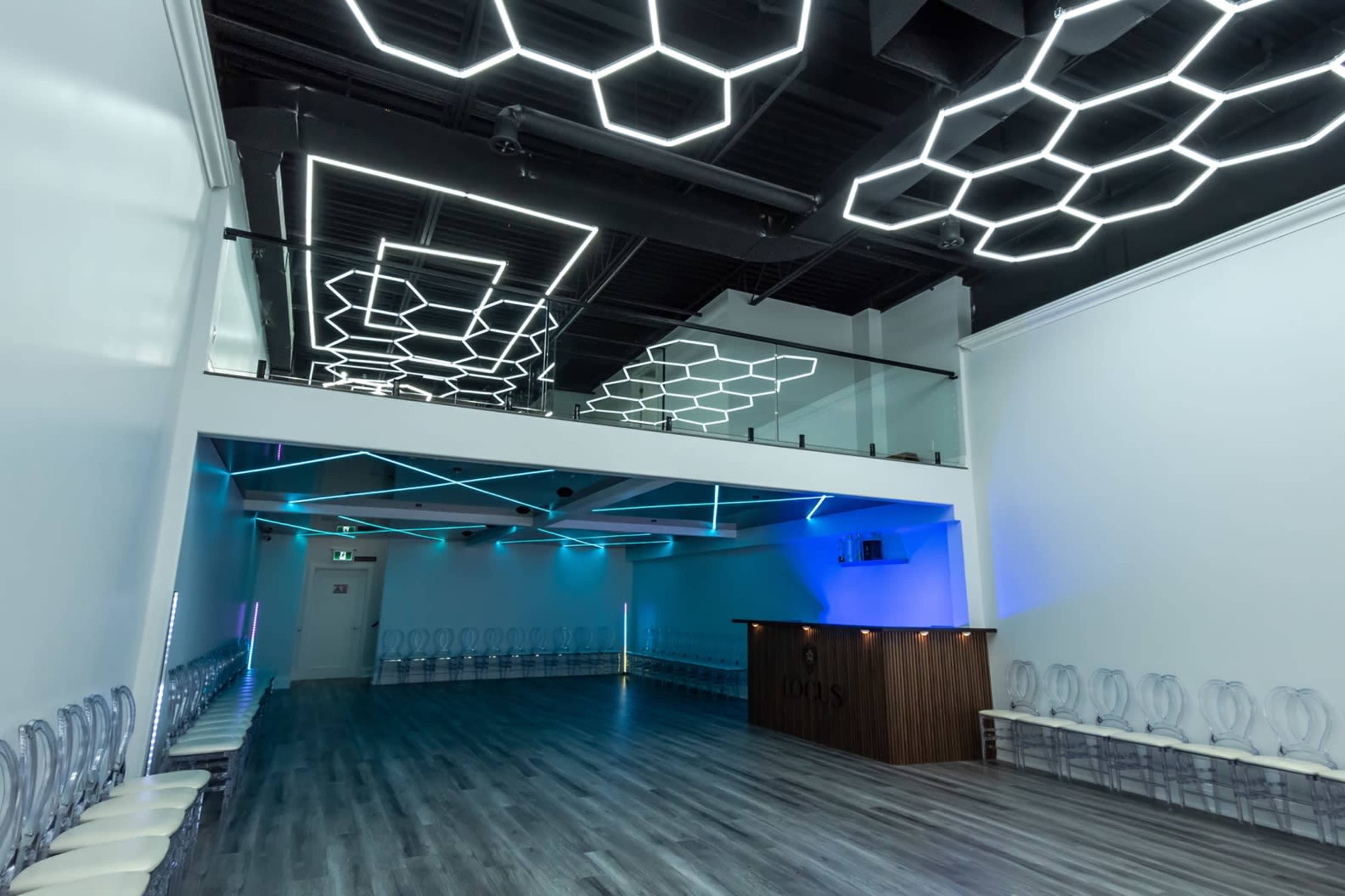 A spacious modern venue with hexagonal light fixtures on the ceiling, a sleek bar counter, and rows of transparent chairs arranged on the floor.