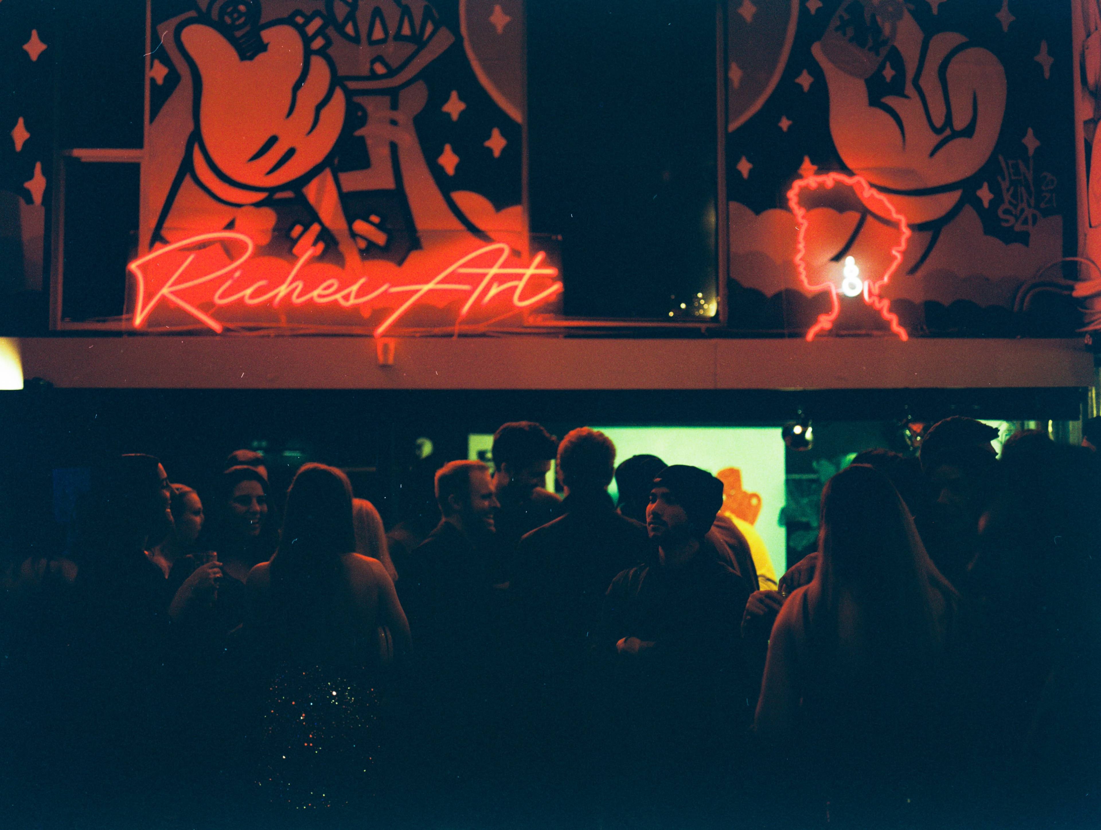 A crowd of people mingles in a dimly lit space featuring vibrant wall art and a glowing neon sign that reads "Riches Art."