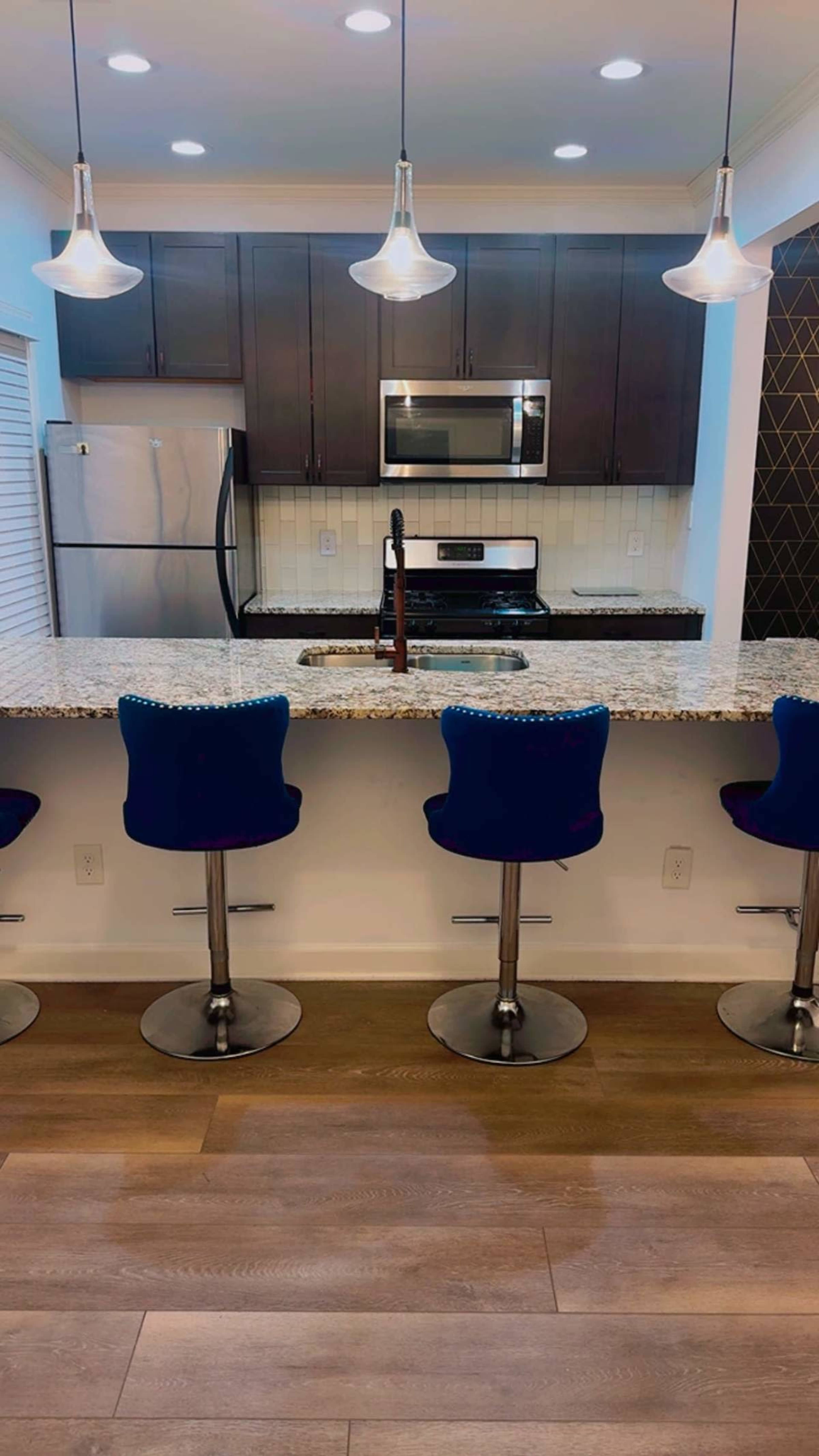 The image shows a modern kitchen with dark cabinetry, stainless steel appliances, and four blue bar stools at a granite countertop.