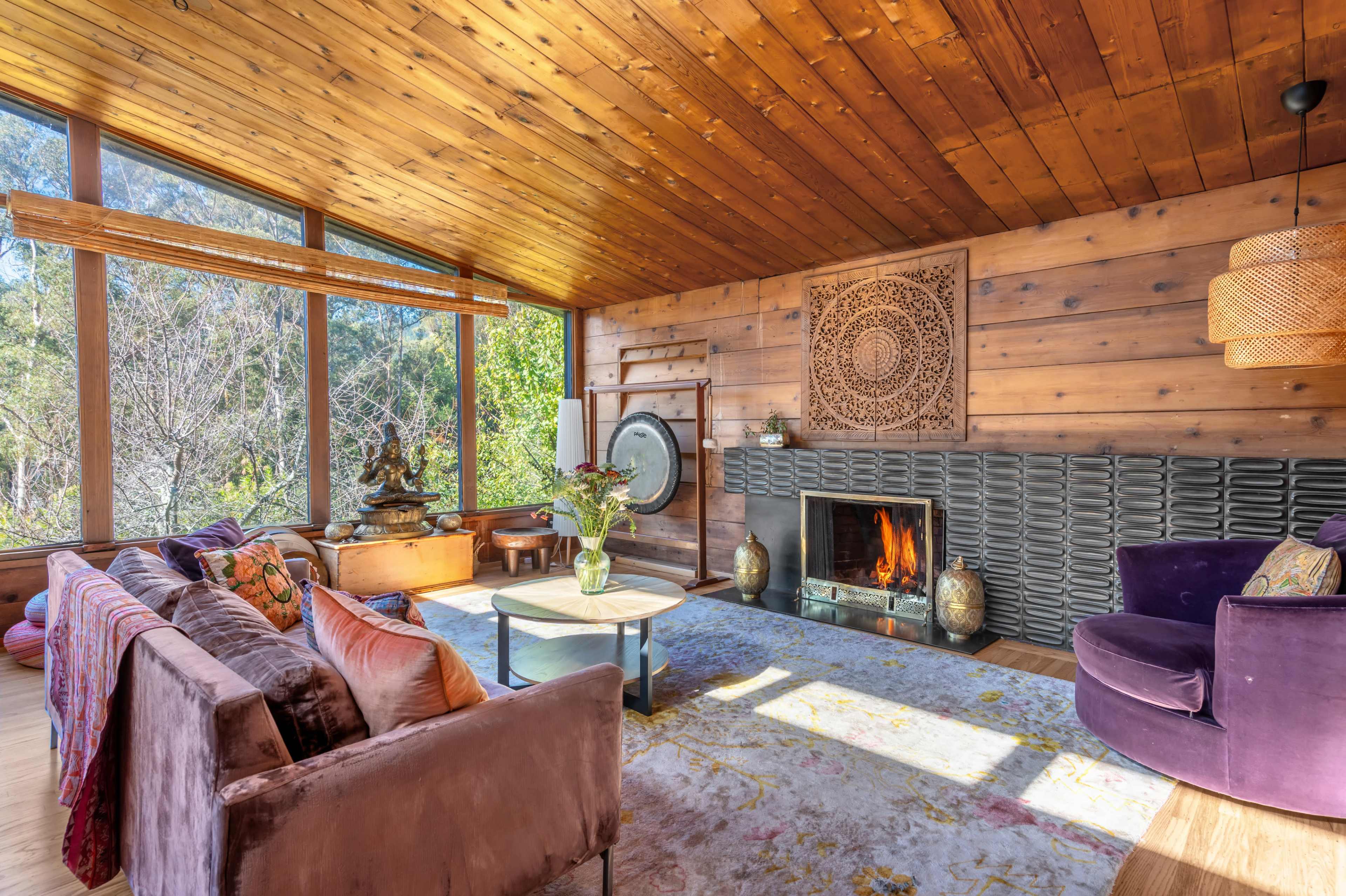 The image depicts a cozy living room with wooden walls, a stone fireplace, a circular coffee table, and large windows offering views of greenery outside.