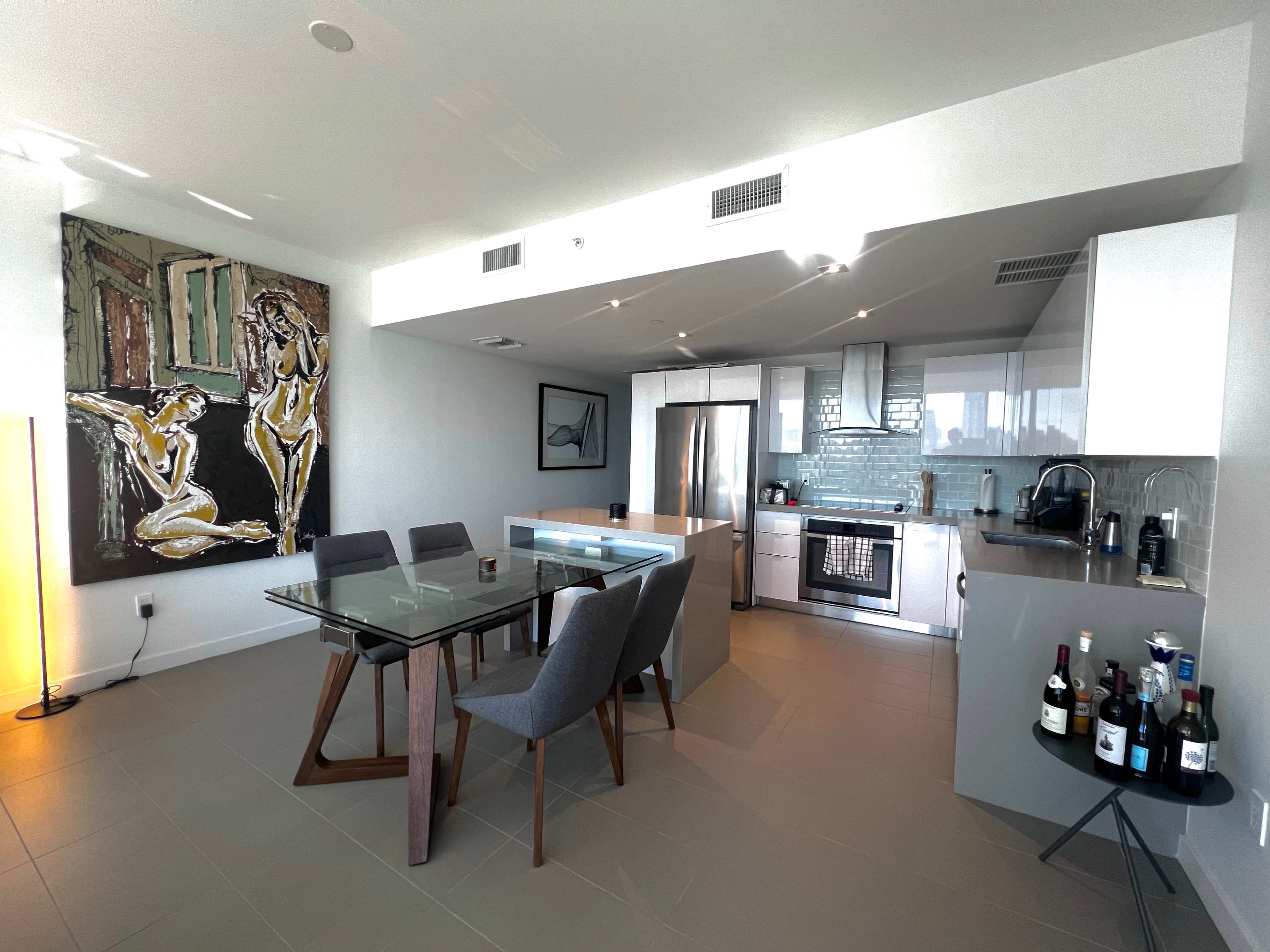 A modern kitchen and dining area feature a glass table with gray chairs, stainless steel appliances, and a large artwork on the wall.