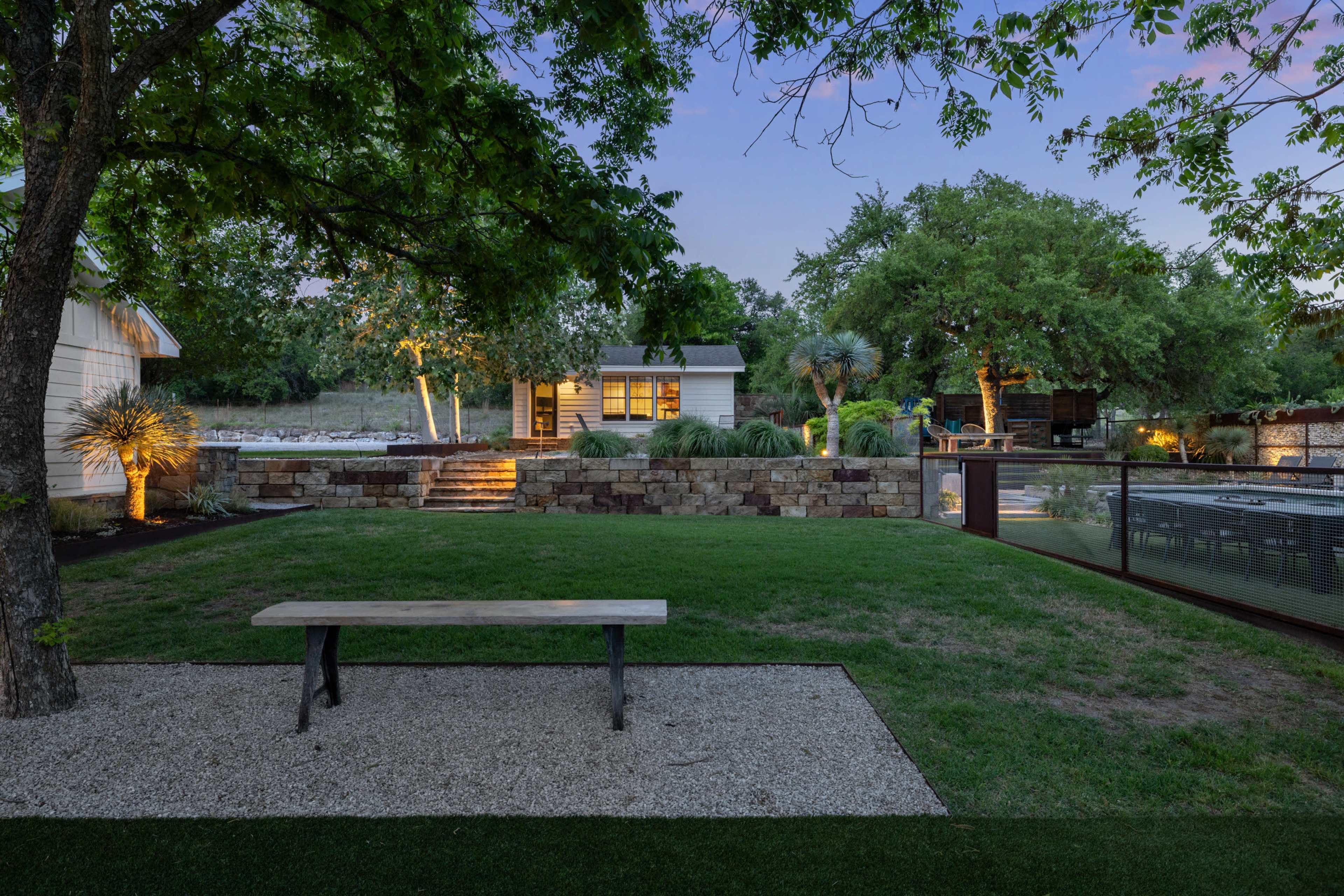 The scene features a landscaped garden with a stone wall, a wooden bench, and a structure in the background, illuminated by soft lighting at dusk.