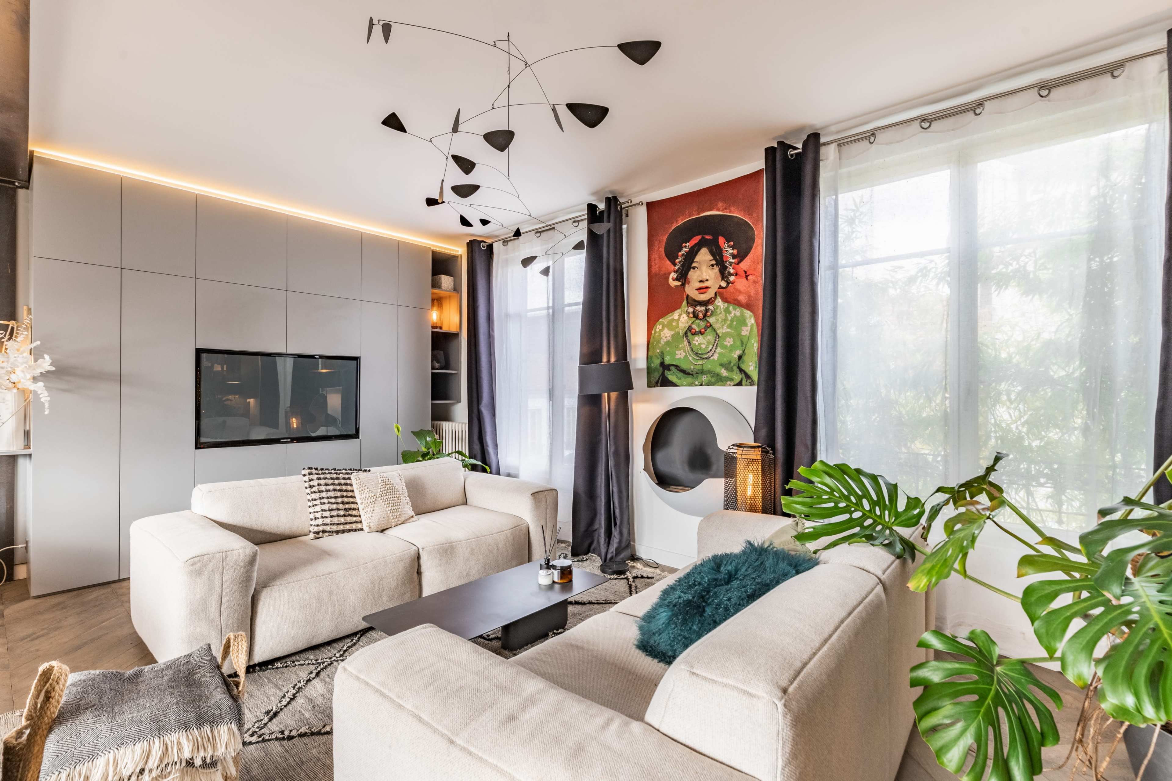 A modern living room features two light-colored sofas, a minimalist black coffee table, a large window with sheer curtains, and an abstract painting on the wall.