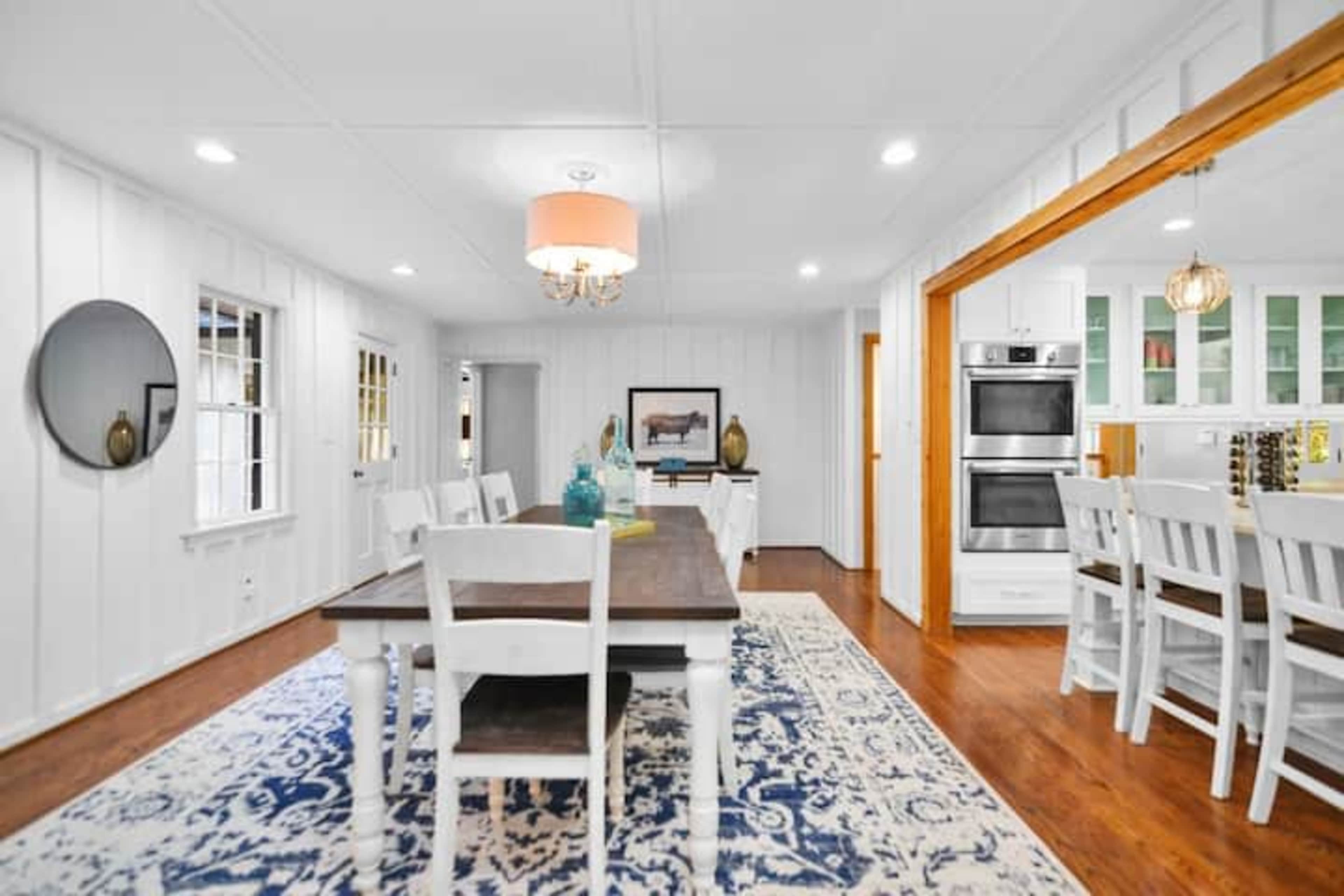 A bright open dining area with a wooden table surrounded by white chairs, a patterned rug, and a view into a kitchen with modern appliances.