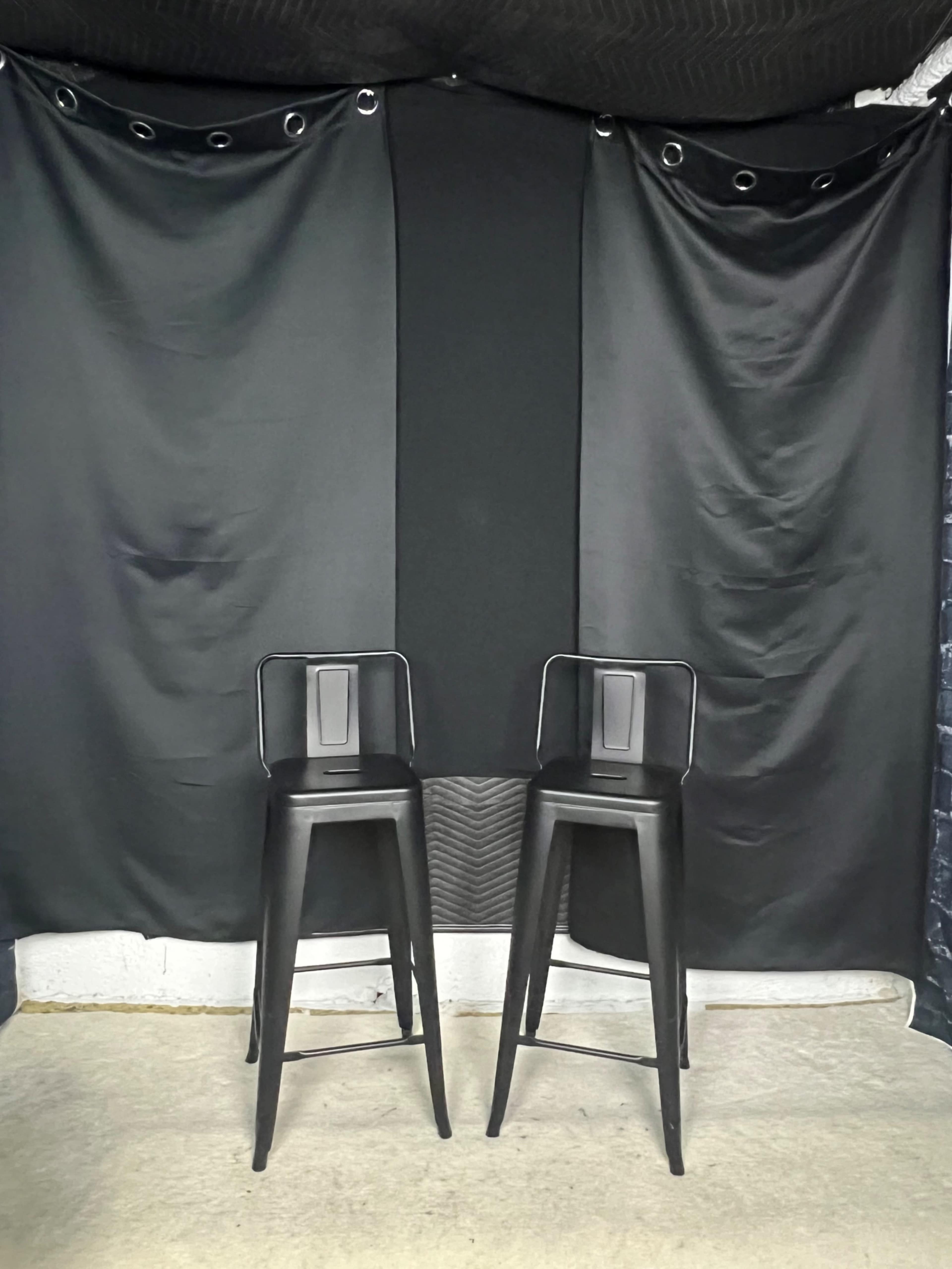 Two black bar stools are positioned in front of a black backdrop with draped fabric.