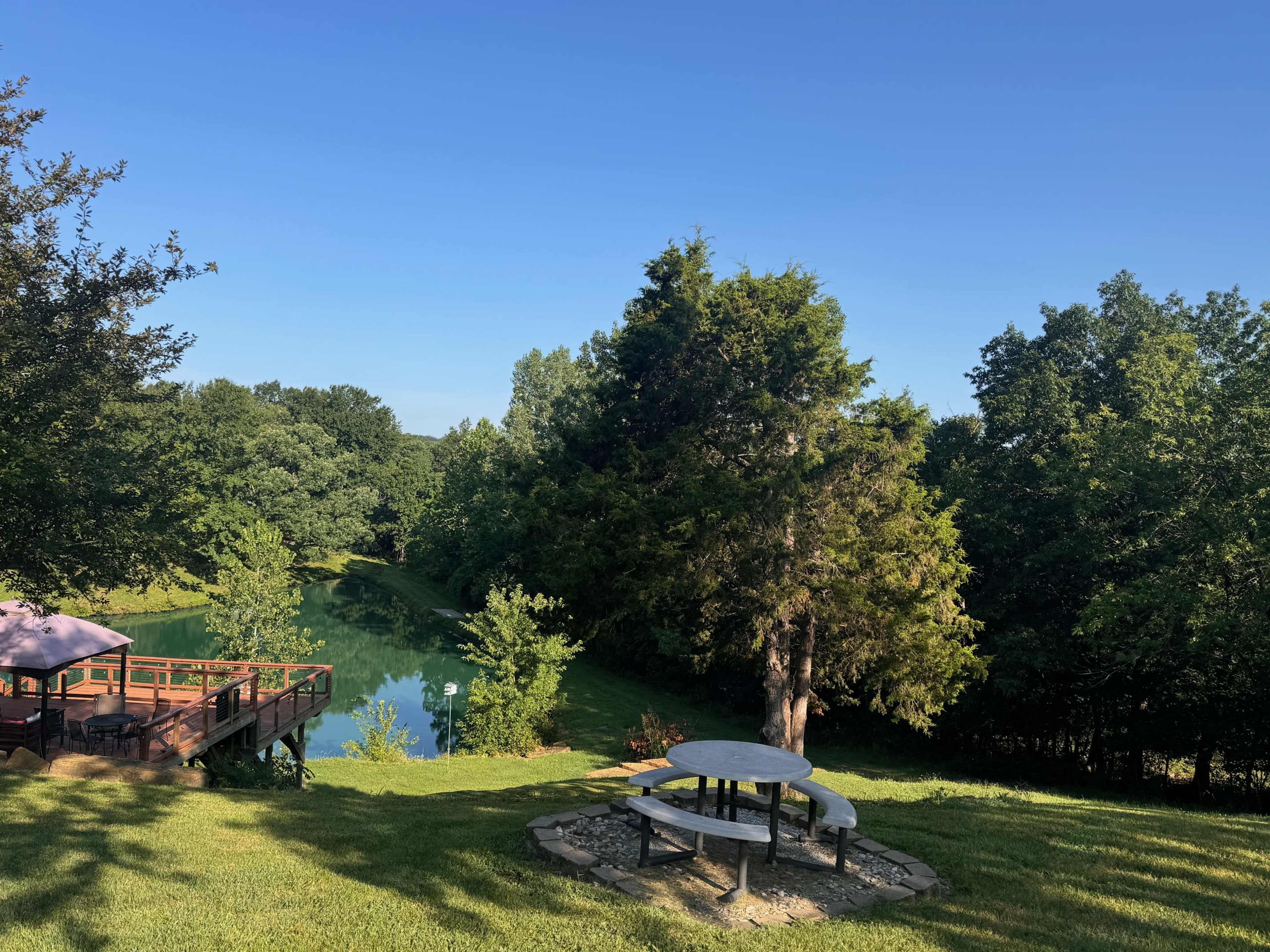 A picnic table is set on a grassy area overlooking a calm pond surrounded by trees.