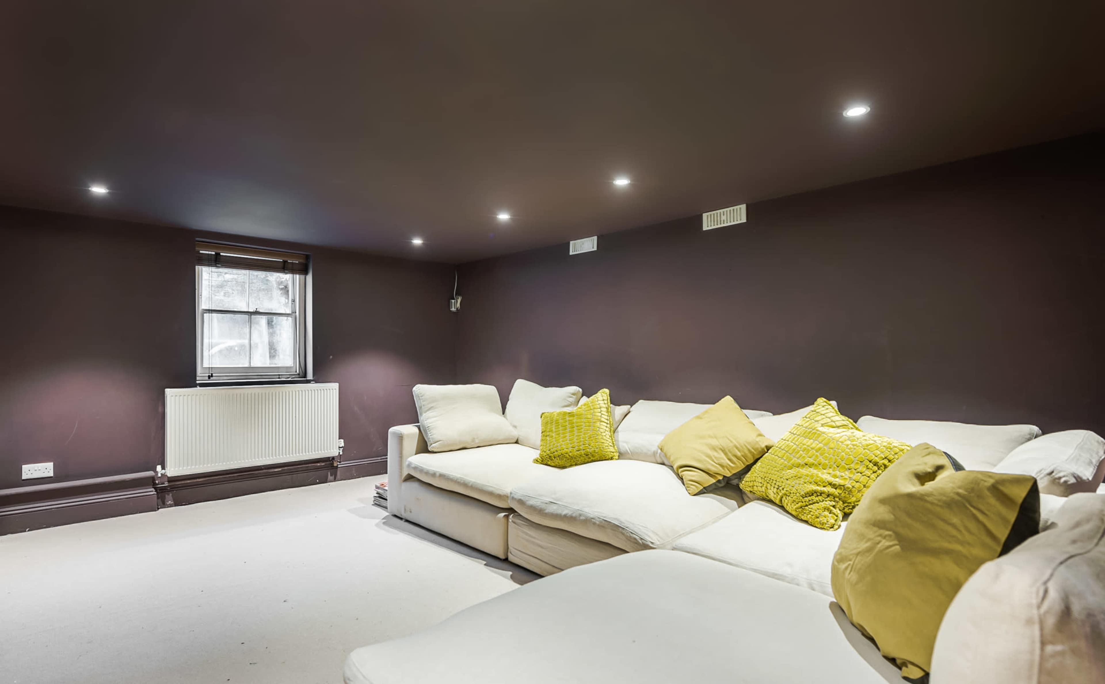 A cozy living room with a large white sectional sofa adorned with yellow pillows, against a dark brown wall and a window.