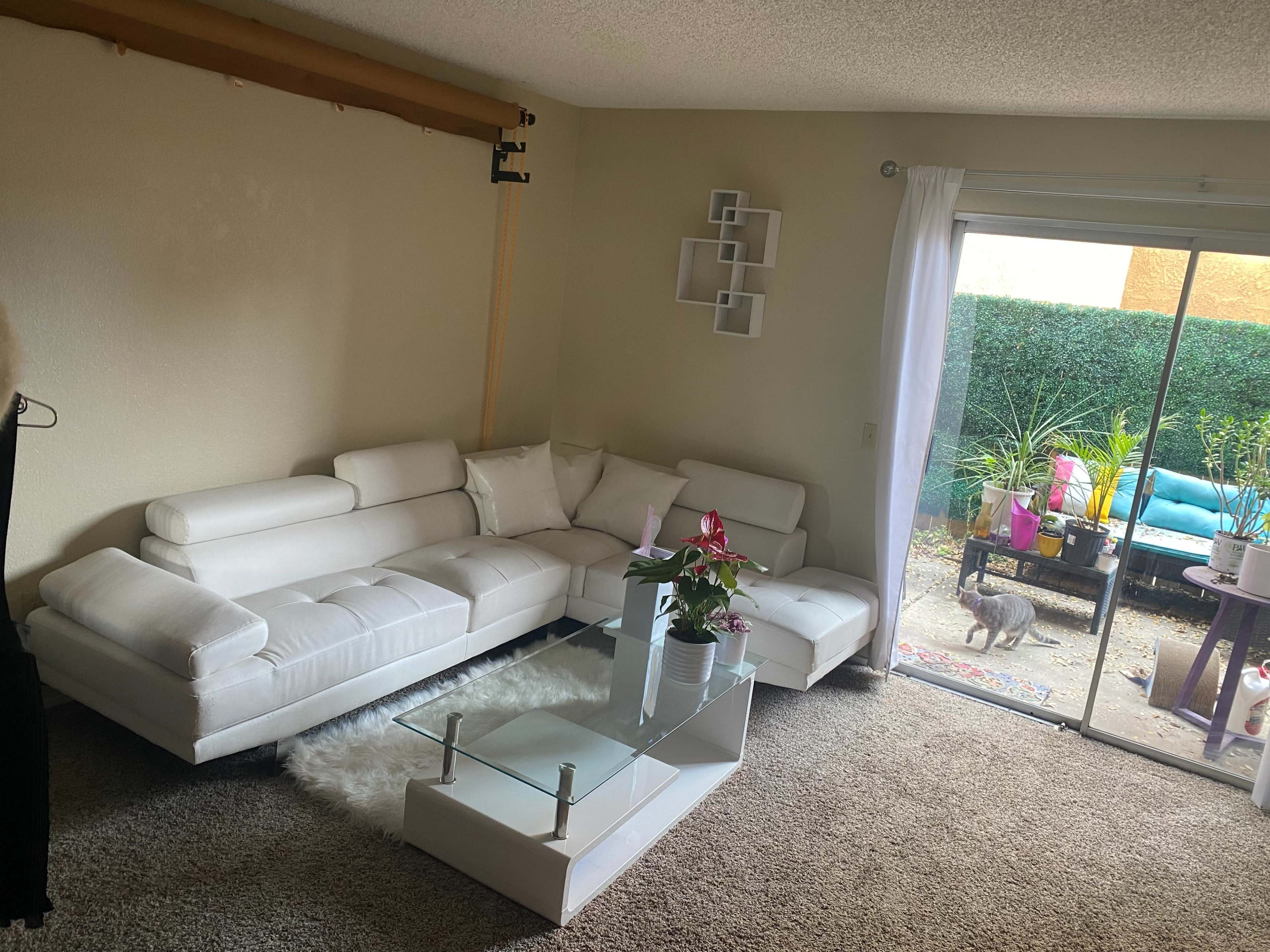 A living room with a white sectional sofa, a glass coffee table, a small plant arrangement, and a sliding door leading to a patio area with outdoor furniture.
