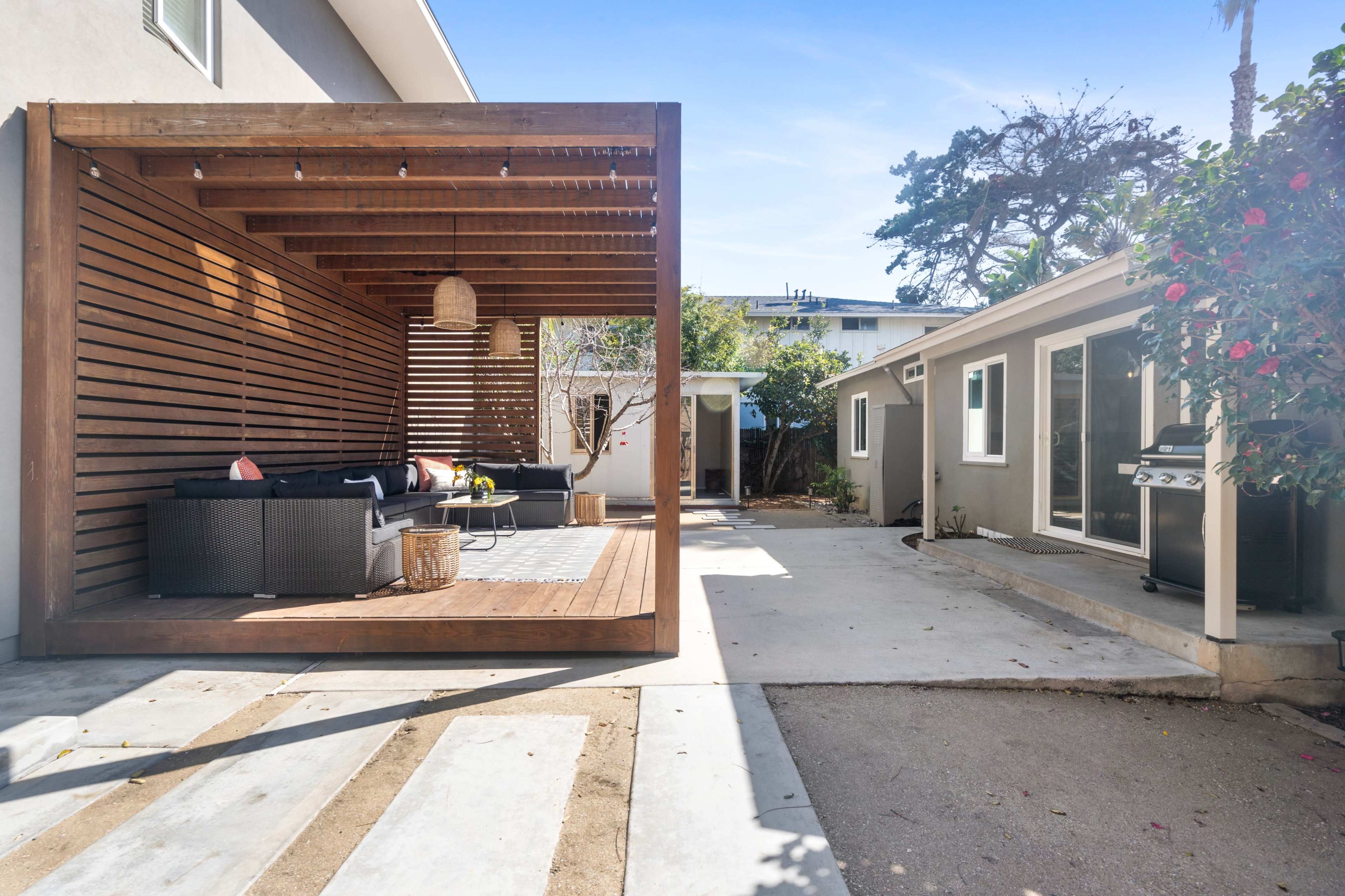 The image shows a modern outdoor patio area with a wooden pergola, featuring a seating arrangement and a grill beside a house.