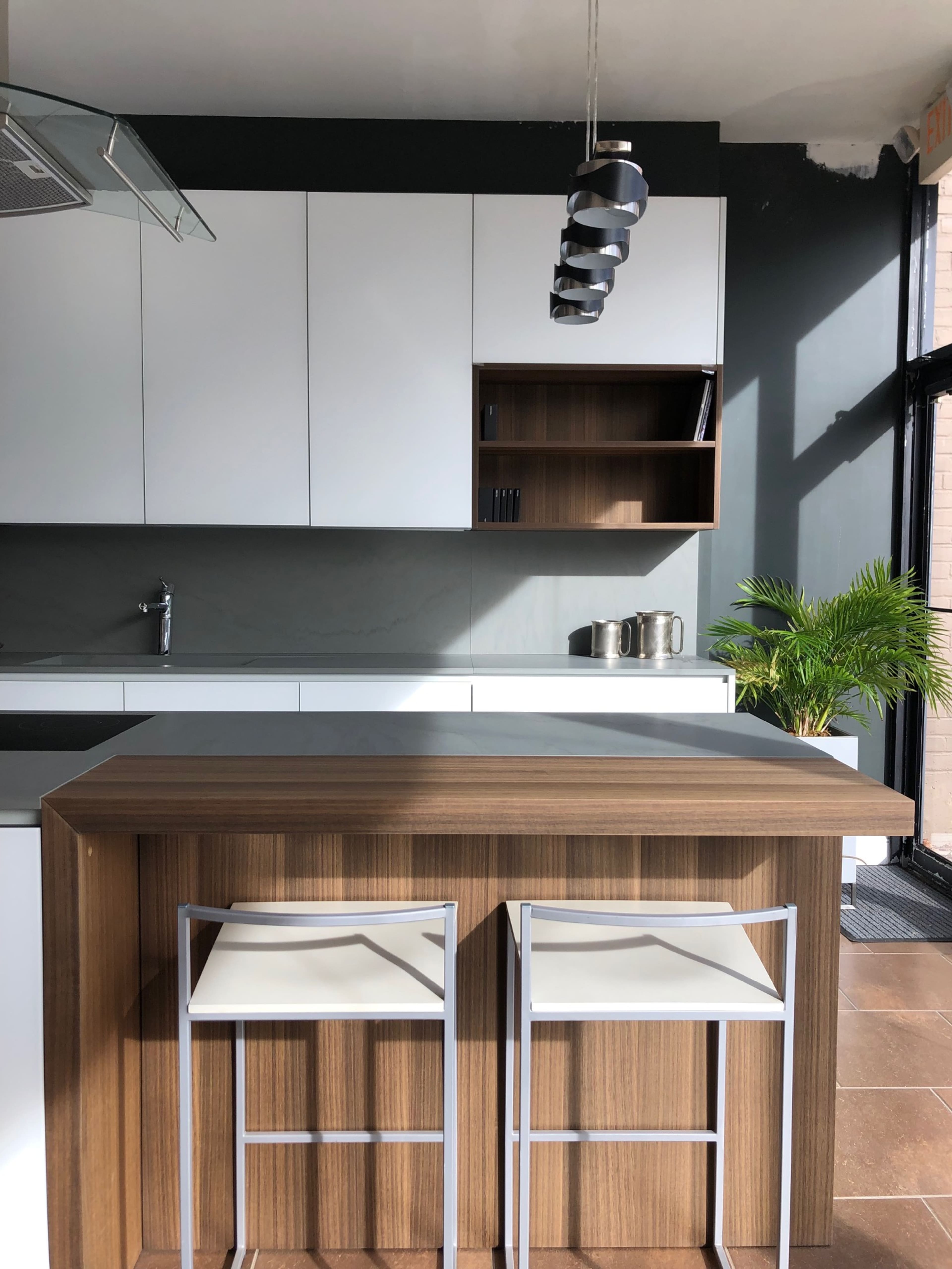 A modern kitchen features a wooden island with two bar stools, sleek white cabinetry, and a large window allowing natural light to illuminate the space.