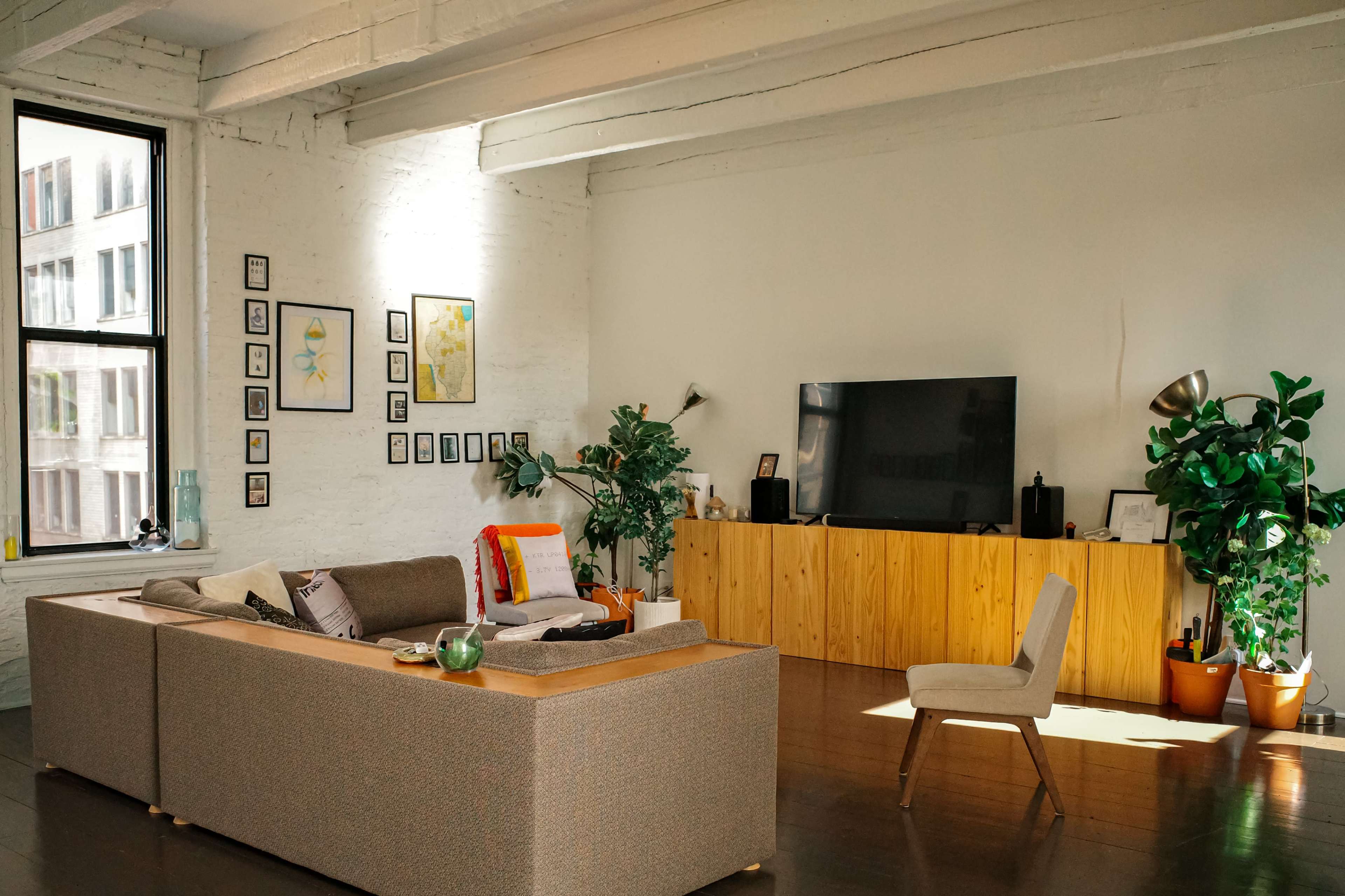 The image shows a modern living room with a large sofa, a wooden TV stand, and decorative plants positioned near windows.