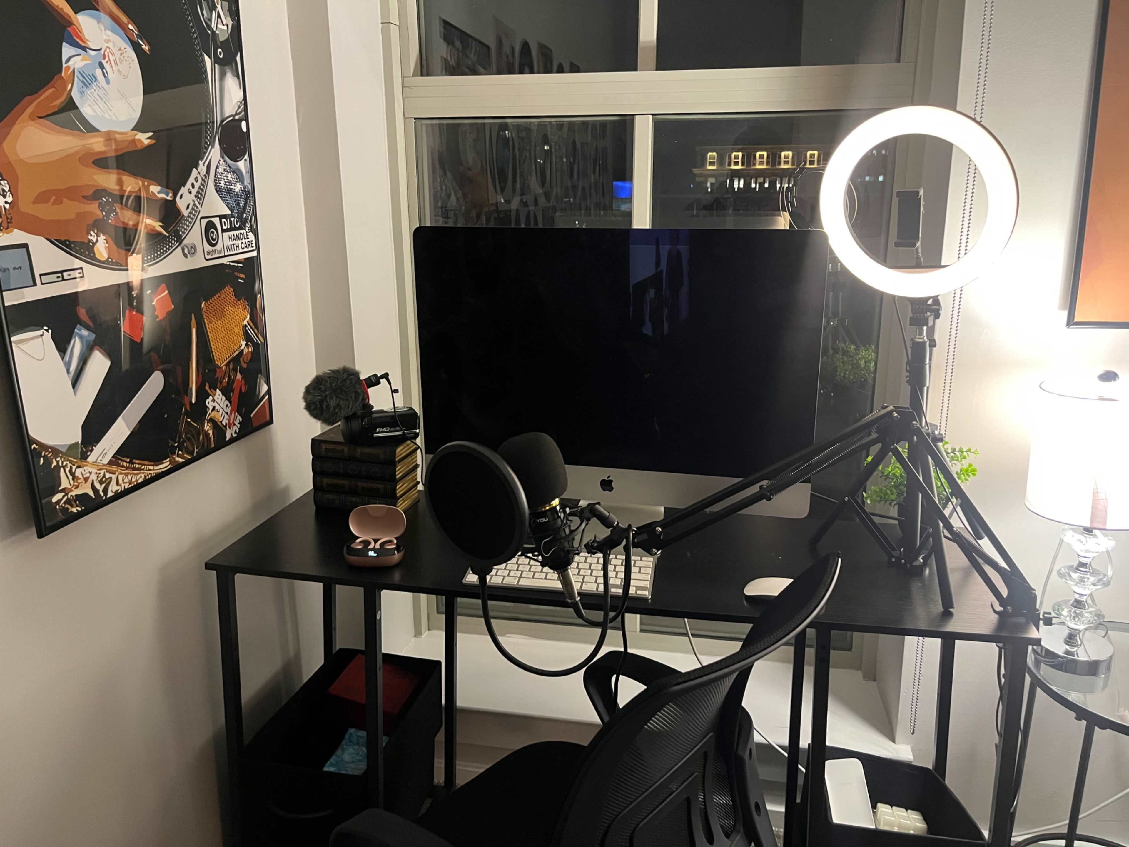 The image shows a modern workspace featuring an iMac on a black desk with a ring light, microphone, and chair, along with decorative items on the wall.