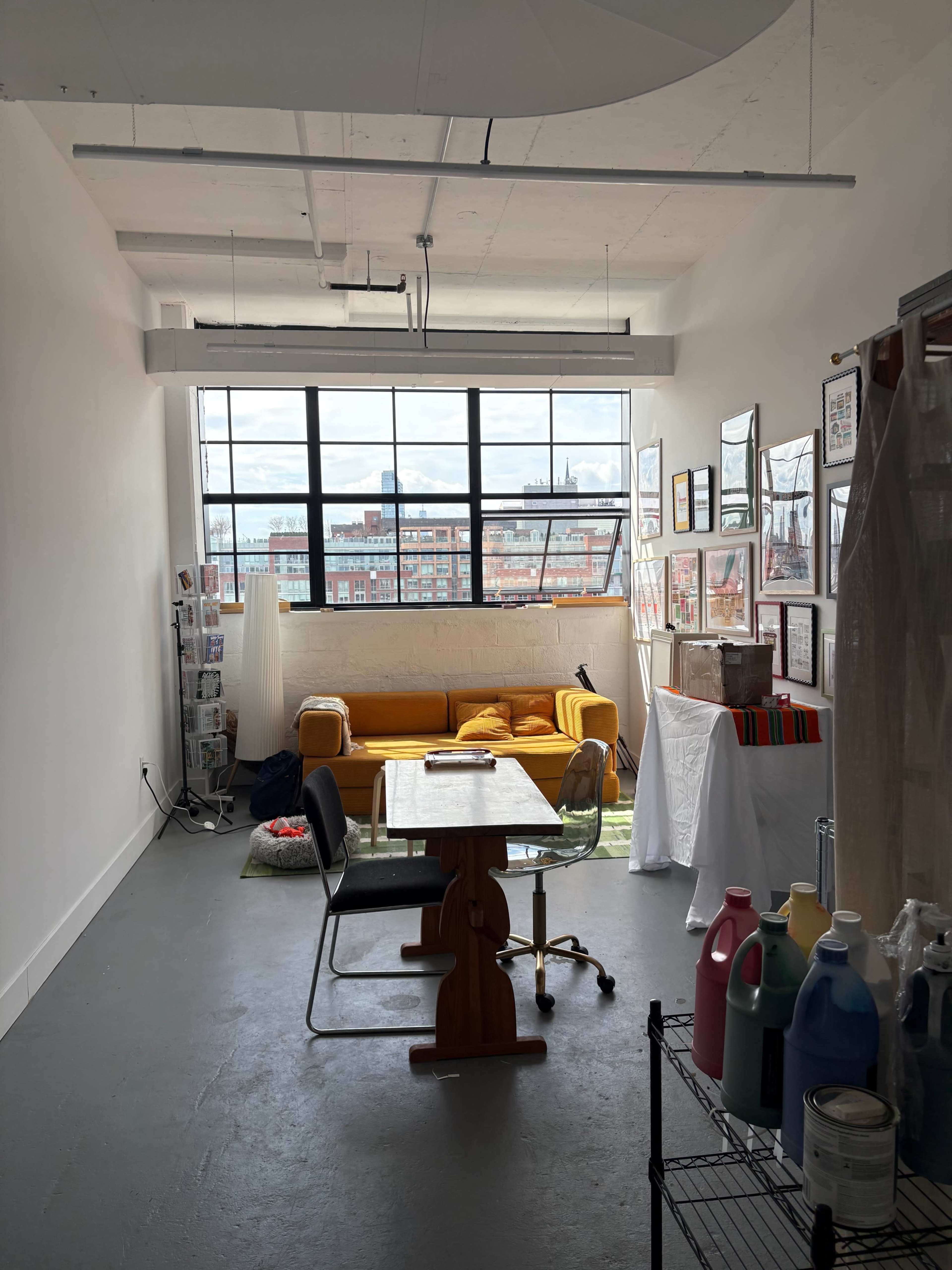The image shows a bright, open studio space with a yellow couch, a wooden table with a chair, and walls adorned with framed artwork, all under a large window overlooking a cityscape.