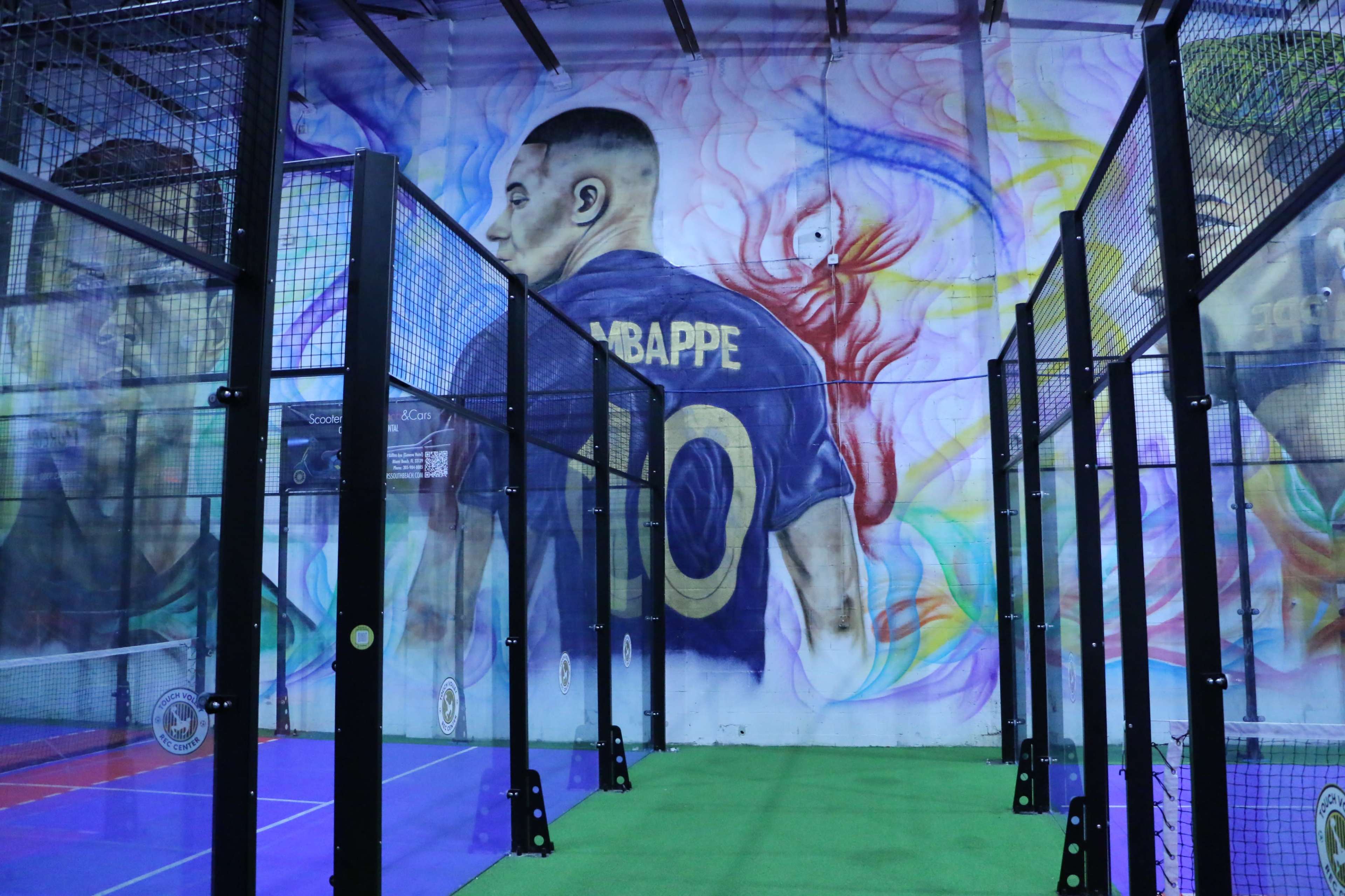 The image shows an indoor sports facility featuring padel courts and a large mural of a player wearing a jersey with the name "MBAPPE" and the number "10" on the wall.