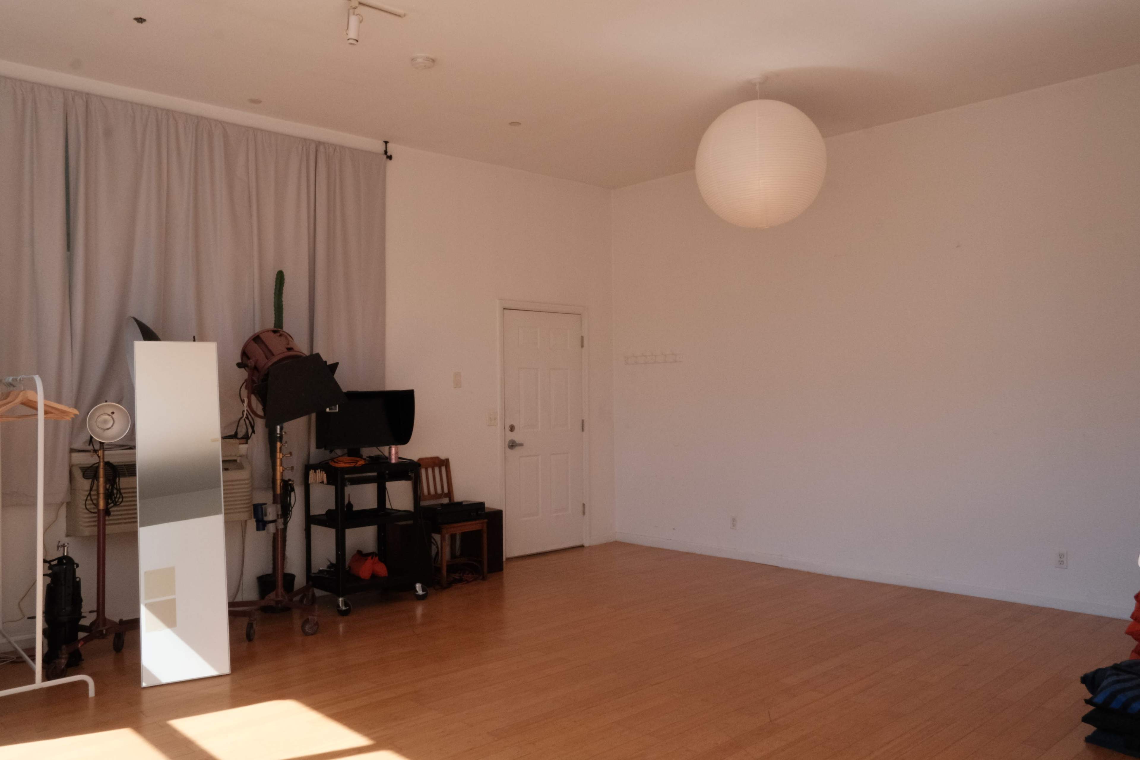 A spacious room with light wooden flooring, a large mirror, a minimalist clothing rack, and a door leading to another area, all illuminated by a paper lantern pendant light.