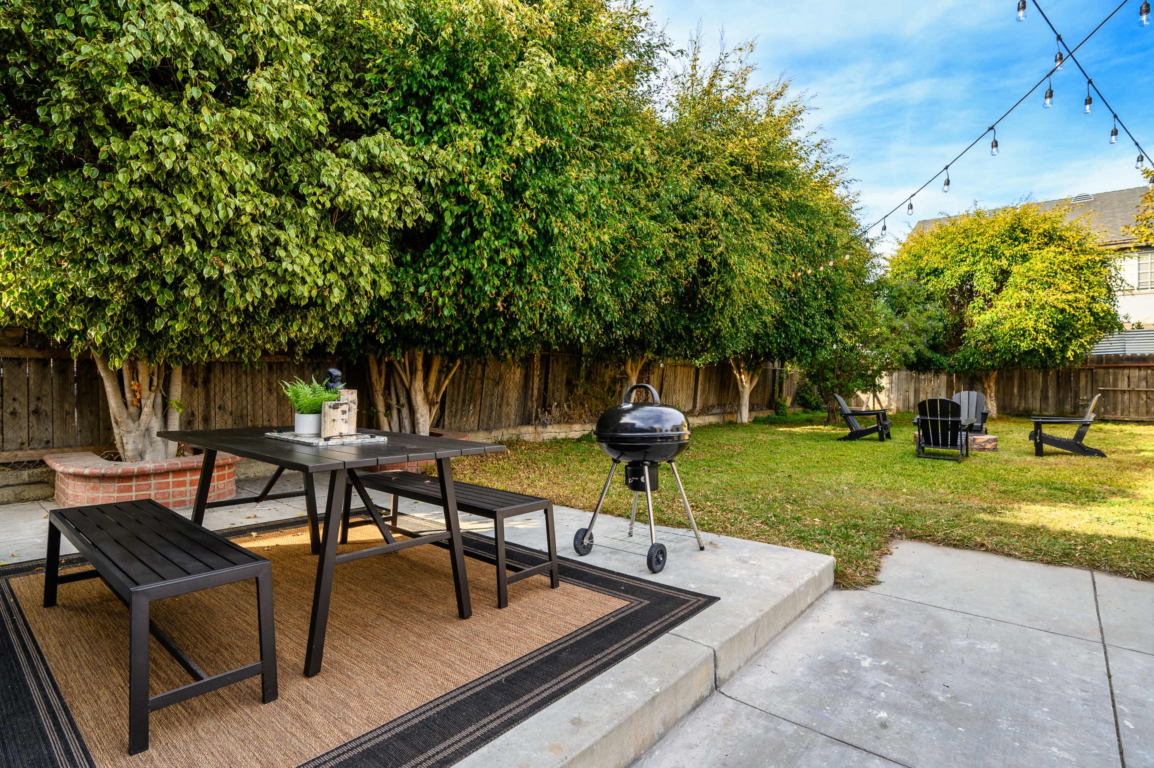 A backyard patio features a black table and benches, a charcoal grill, and several trees, with string lights overhead and a grassy area nearby.
