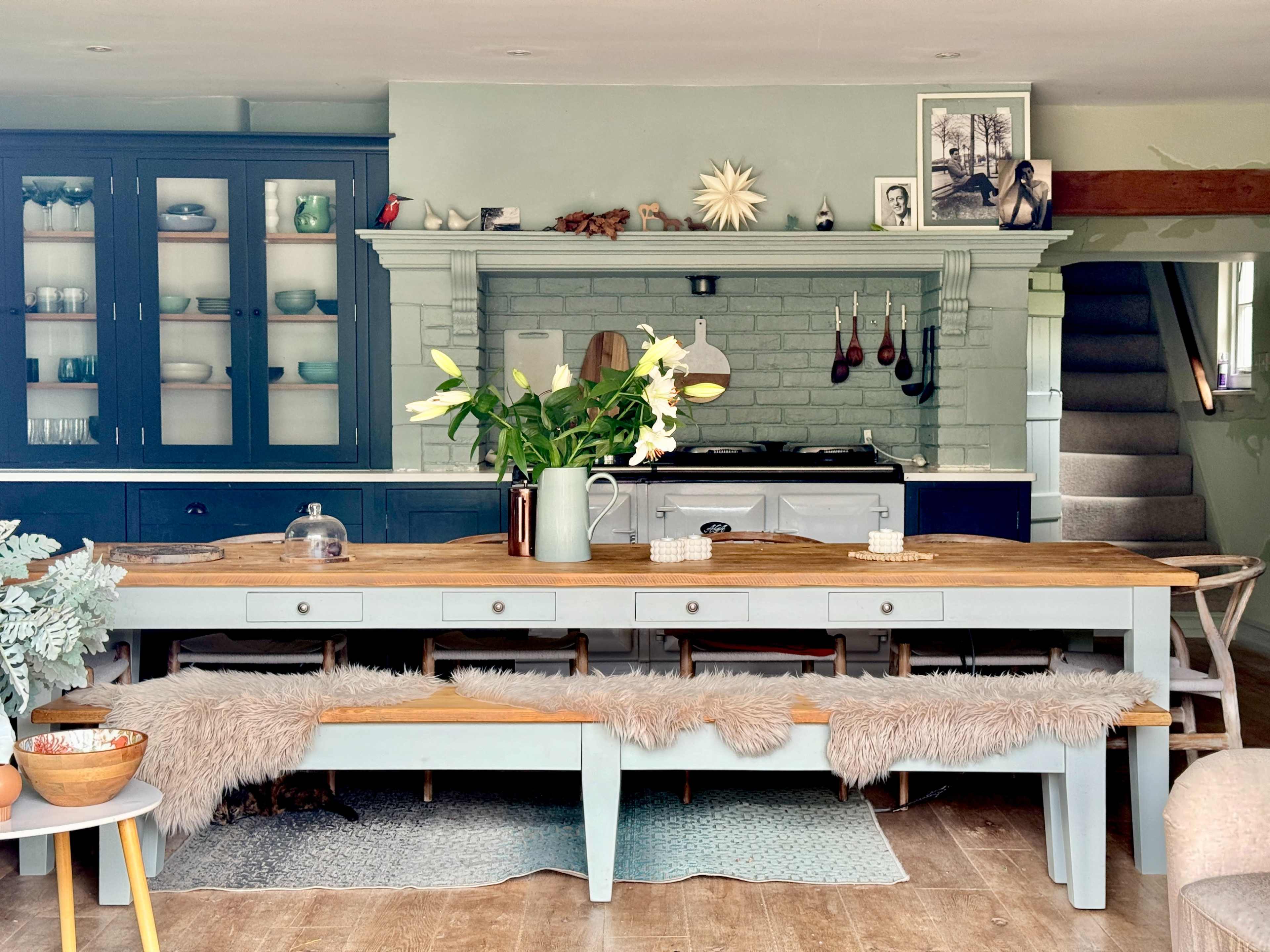 A spacious kitchen featuring a large wooden dining table with benches, surrounded by a blue cabinetry unit and a white stove, accented by decorative plants and art.