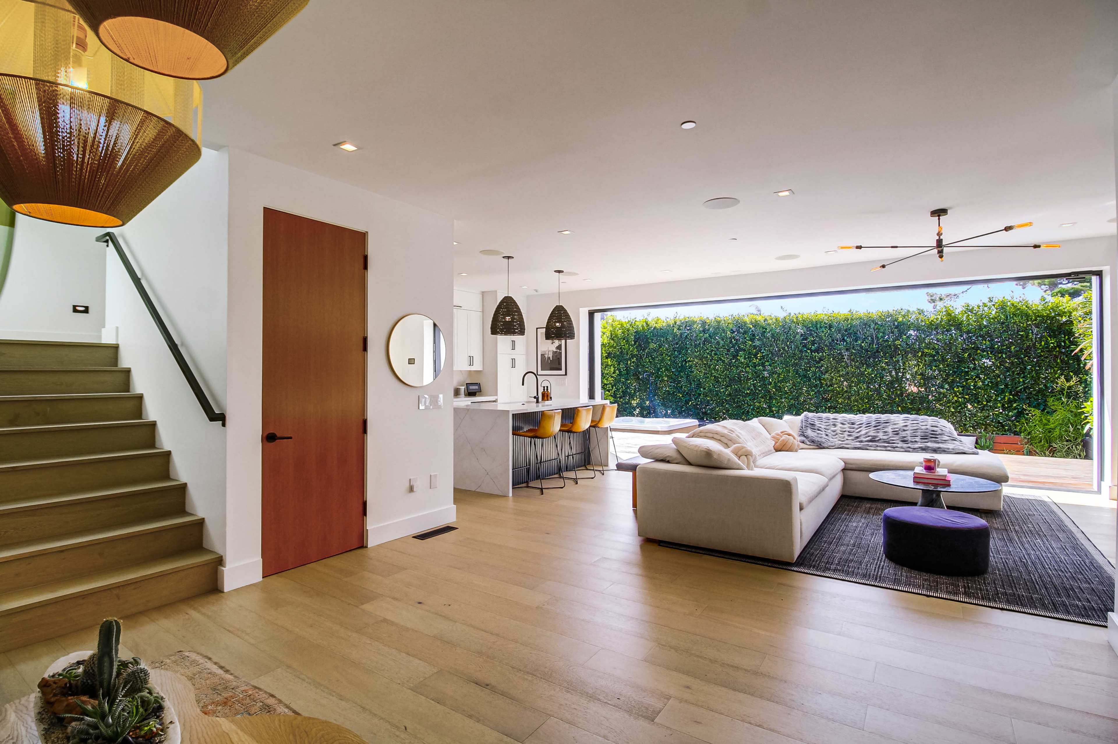 The image shows a modern living space featuring a white sectional sofa, a dining area with bar stools, and large windows overlooking a green hedge.