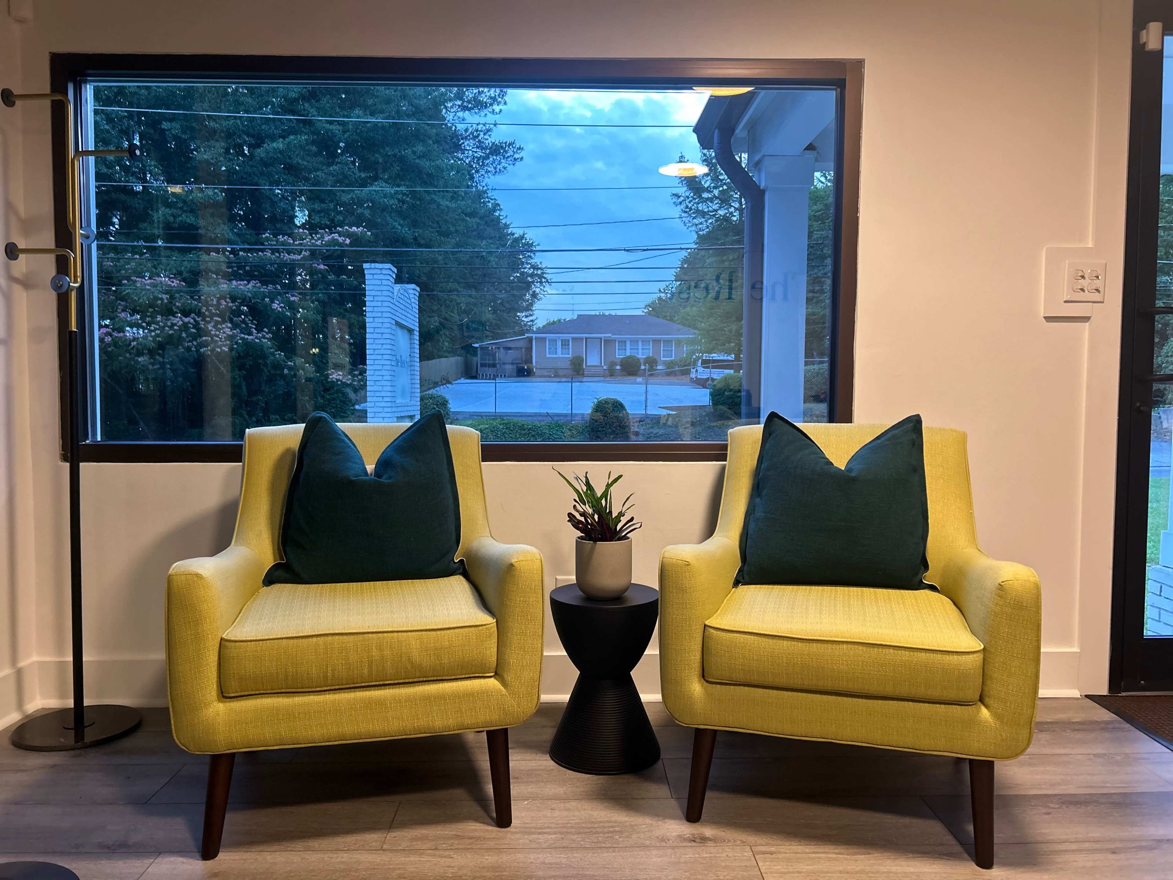 Two yellow chairs with green pillows are positioned facing a window that overlooks a street lined with trees.