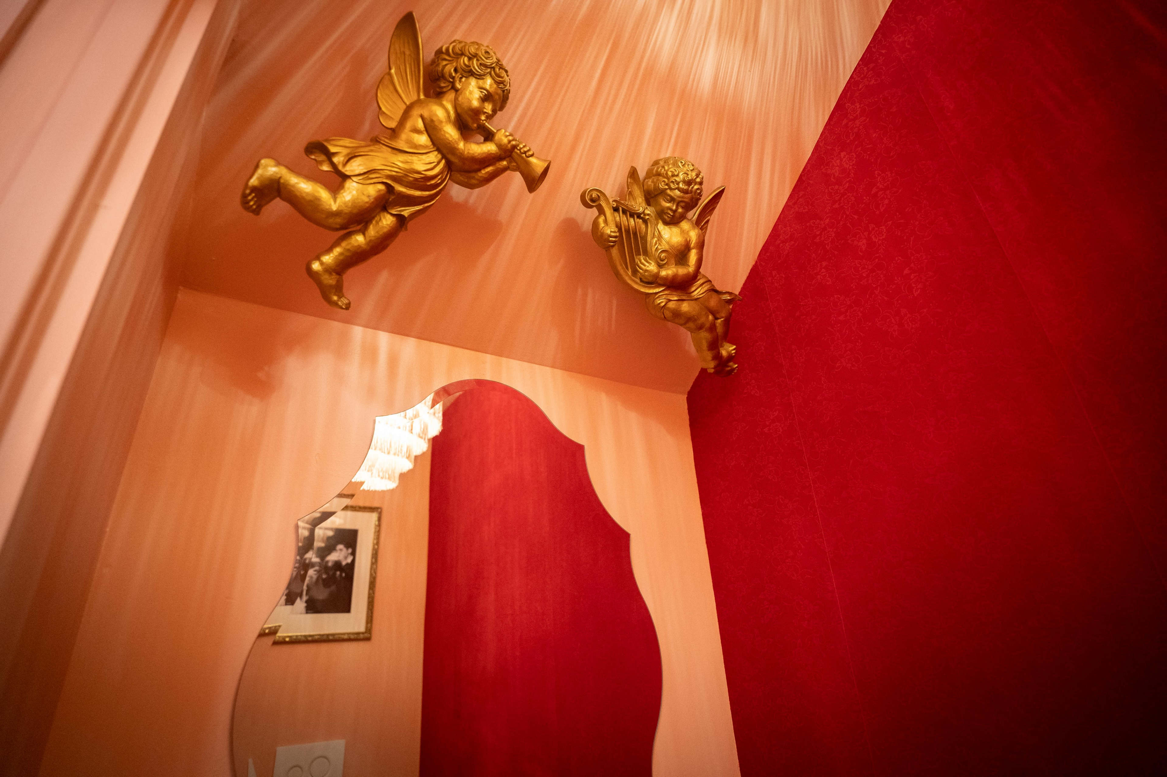 The image features a corner of a room with golden cherubs hanging from the ceiling and a large mirror reflecting the surroundings.
