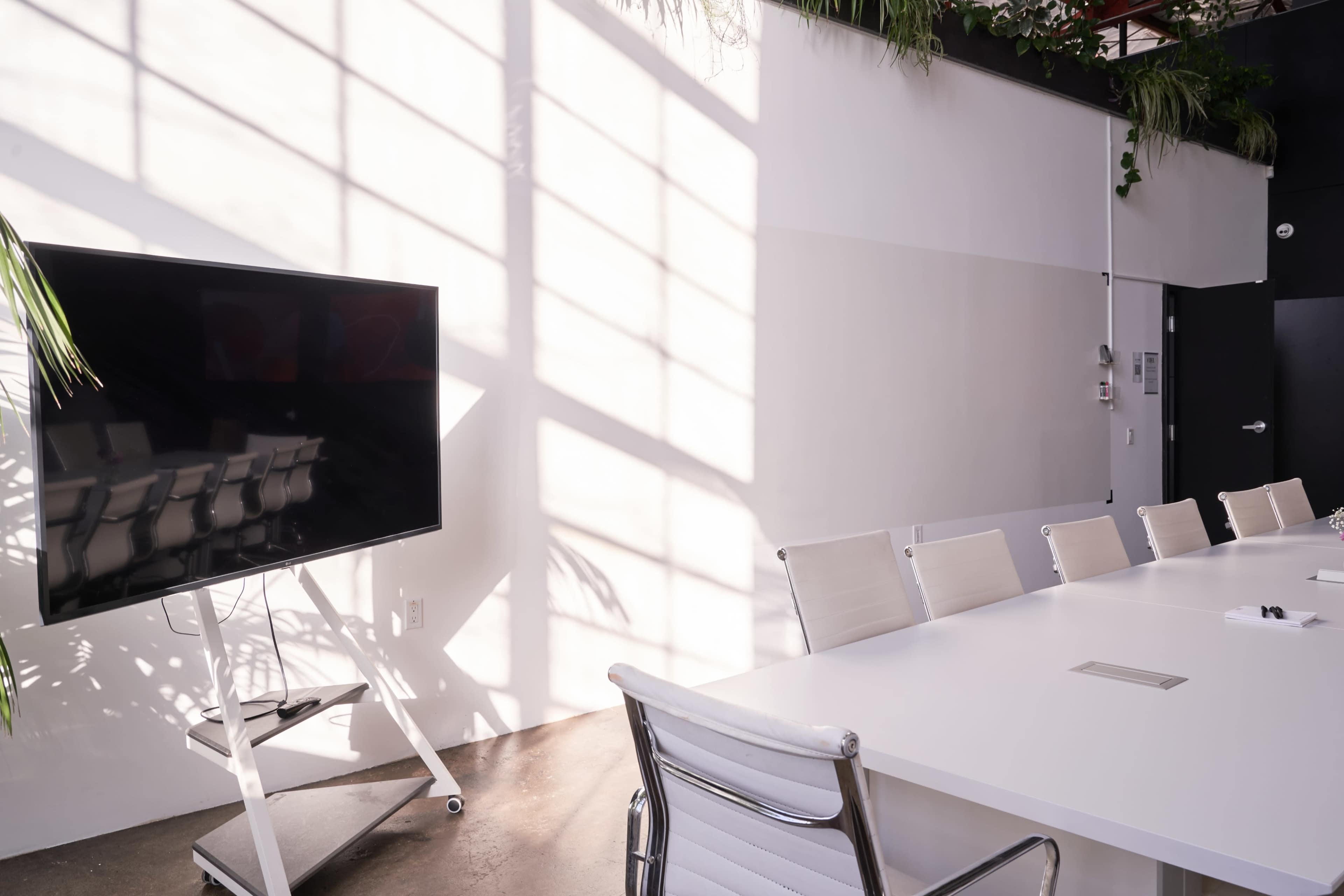 A modern meeting room features a large white table surrounded by silver chairs, with a wall-mounted TV and sunlight casting shadows across the space.