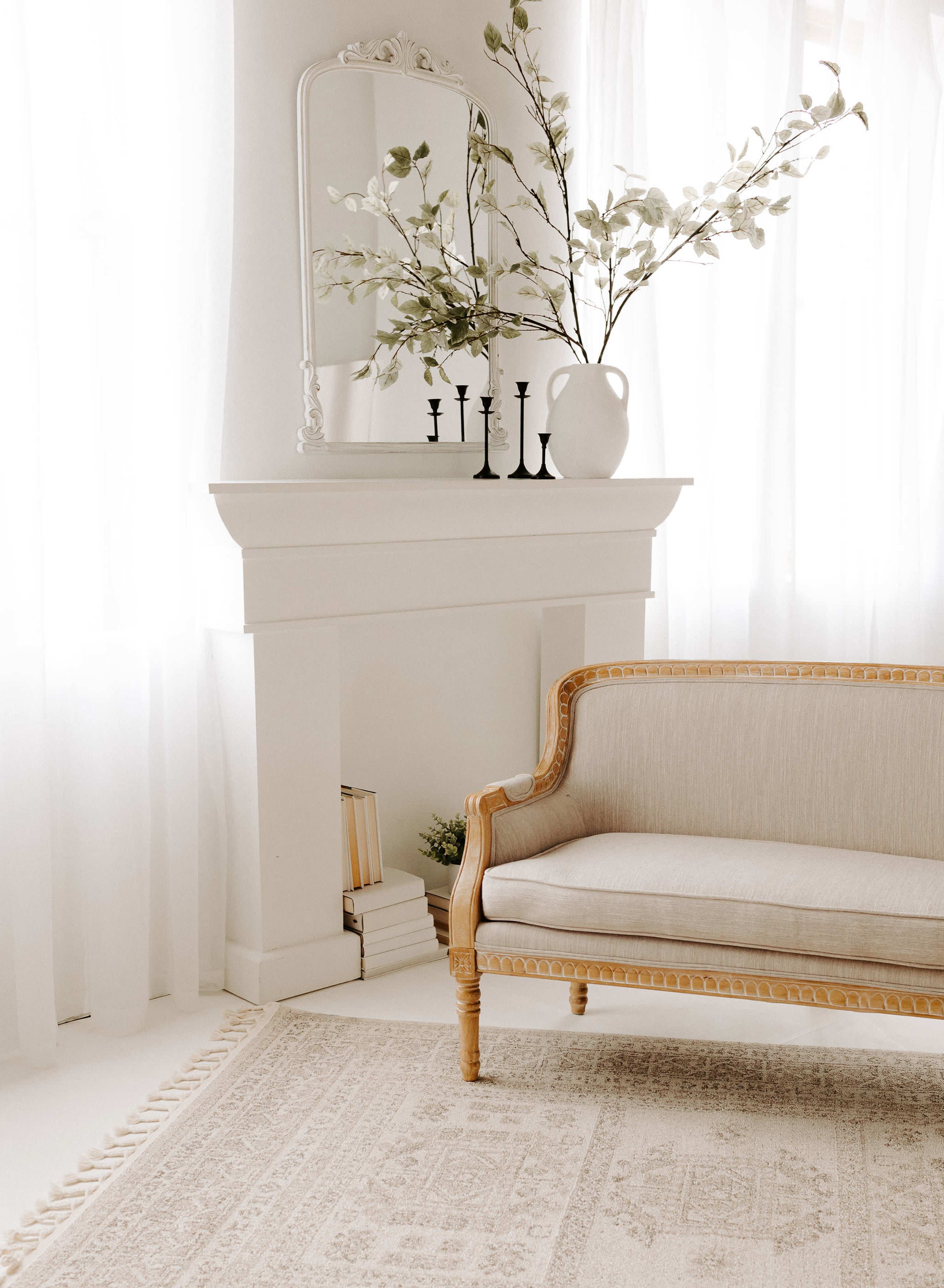 A light-filled room features a white mantel adorned with a vase of greenery, candles, and a mirror, alongside a beige upholstered sofa against a textured rug.