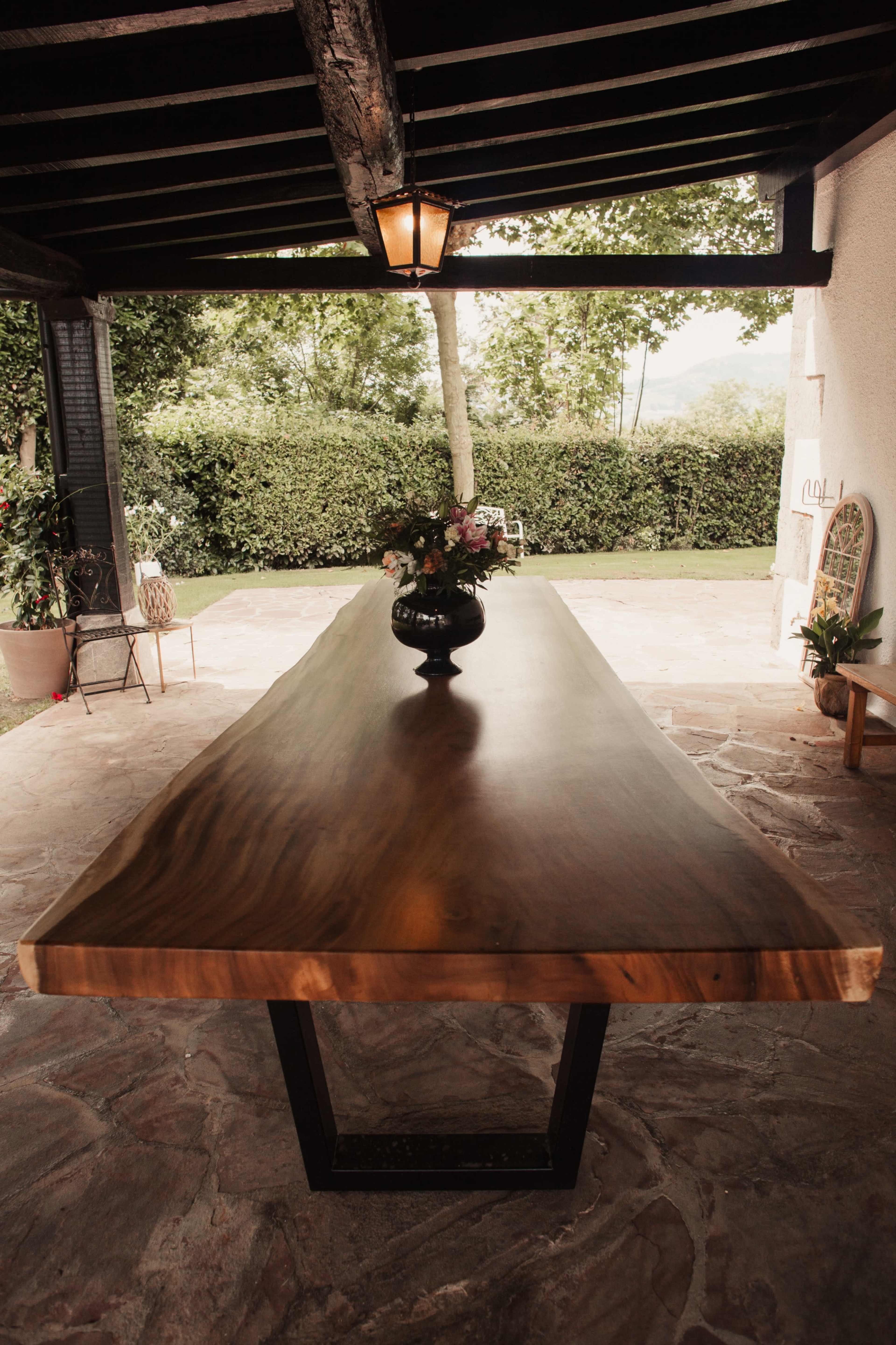 A long, wooden dining table with a natural edge is placed on a stone patio, surrounded by greenery and a decorative vase of flowers.