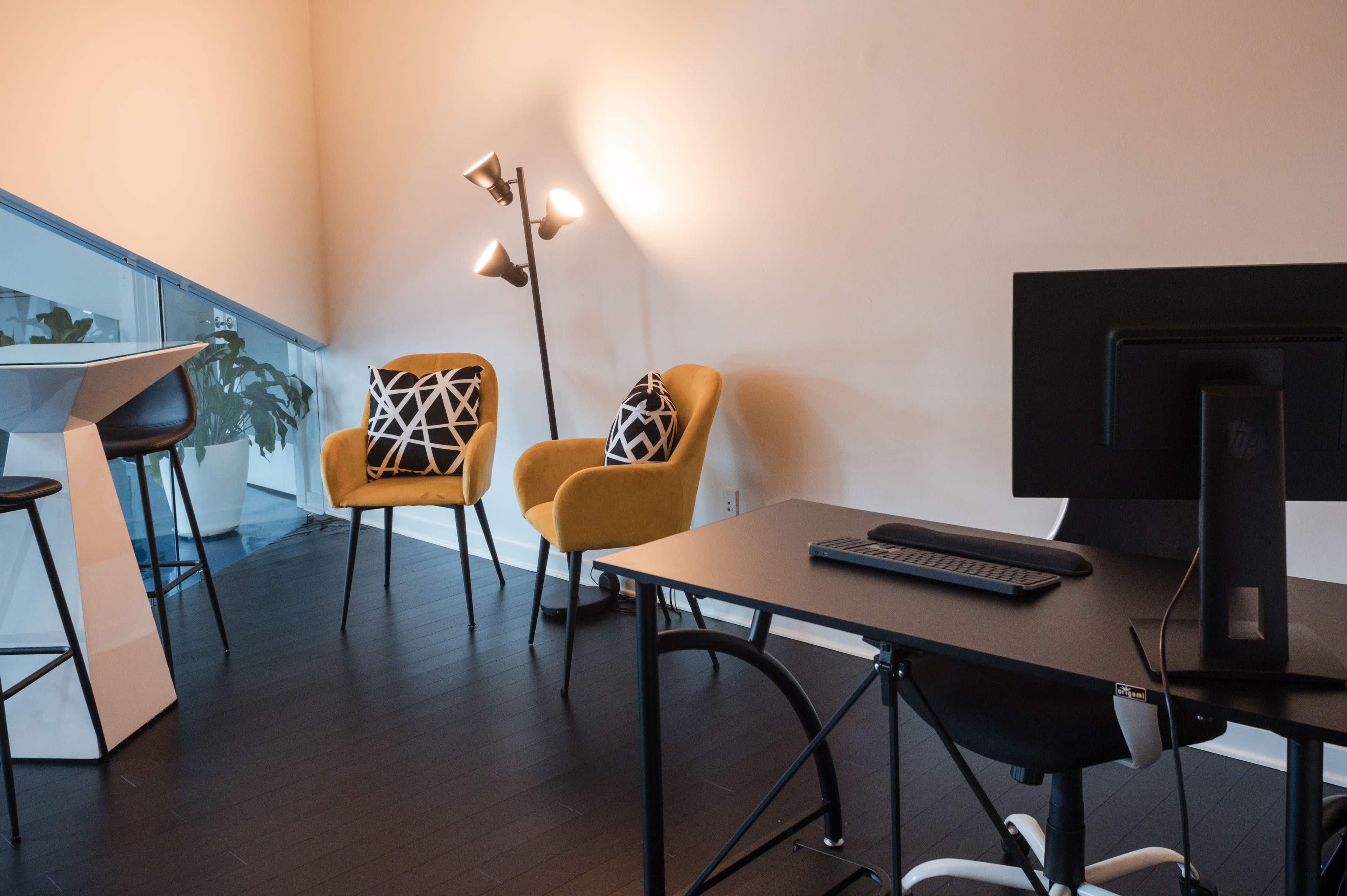 A modern workspace features a black desk with a monitor and keyboard, two yellow chairs with patterned pillows, a standing lamp, and a tall table.