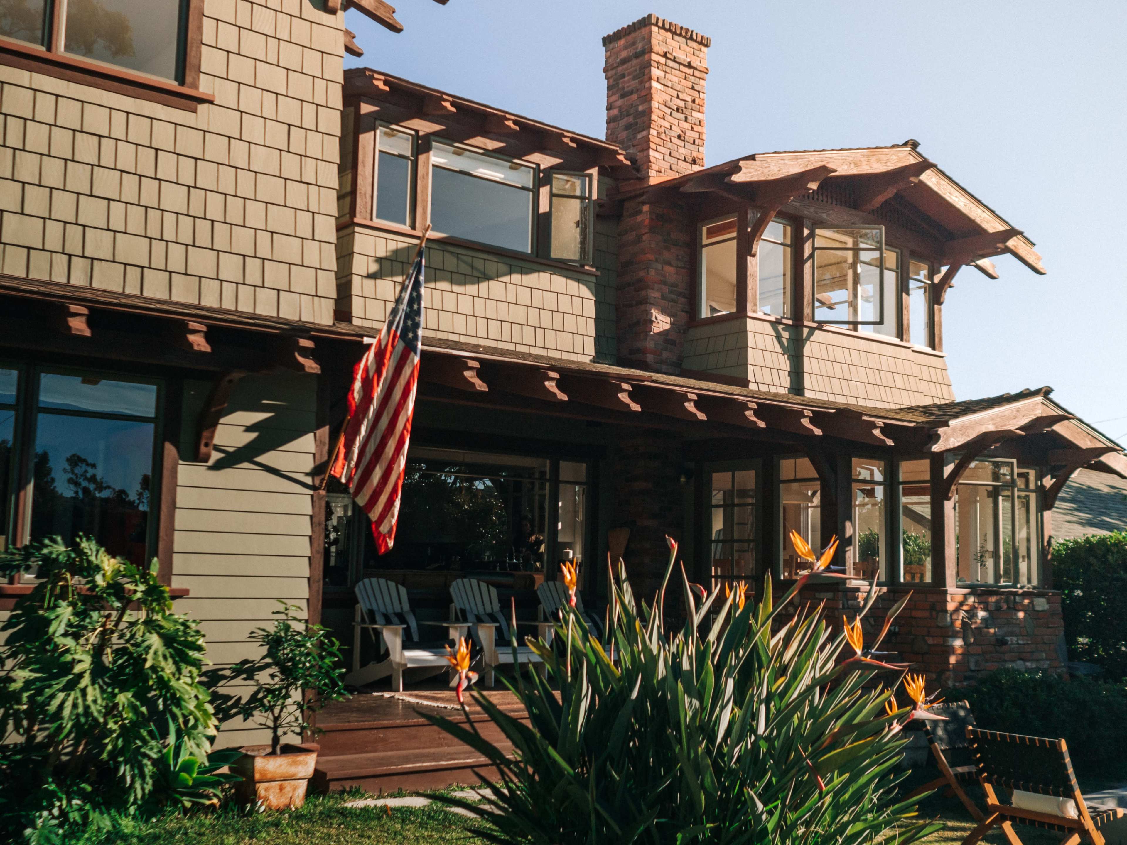 A two-story house with a brick chimney and an American flag, surrounded by tropical plants and large windows.