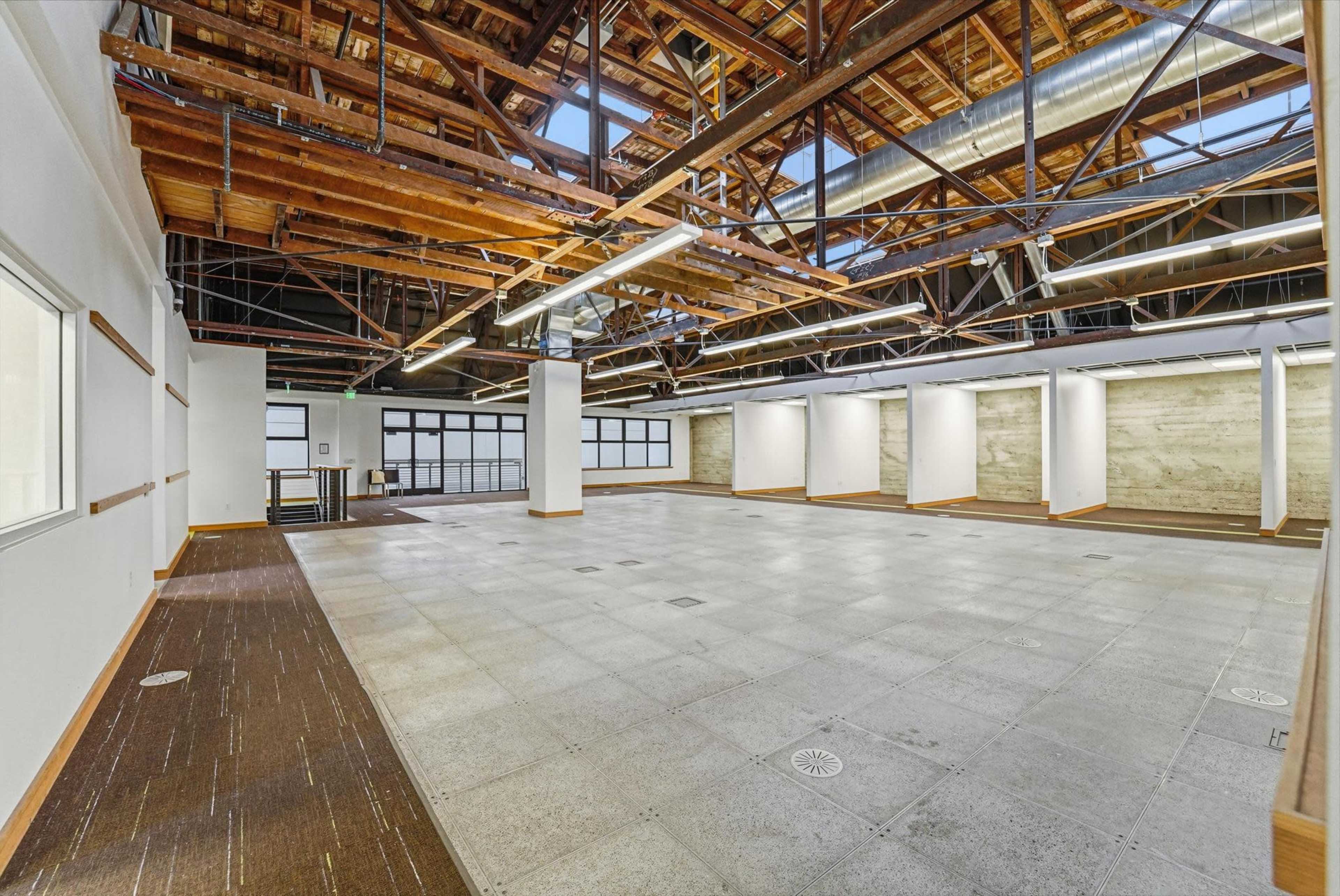 The image shows a spacious, open interior room with high ceilings featuring exposed wooden beams and a mix of flooring materials.