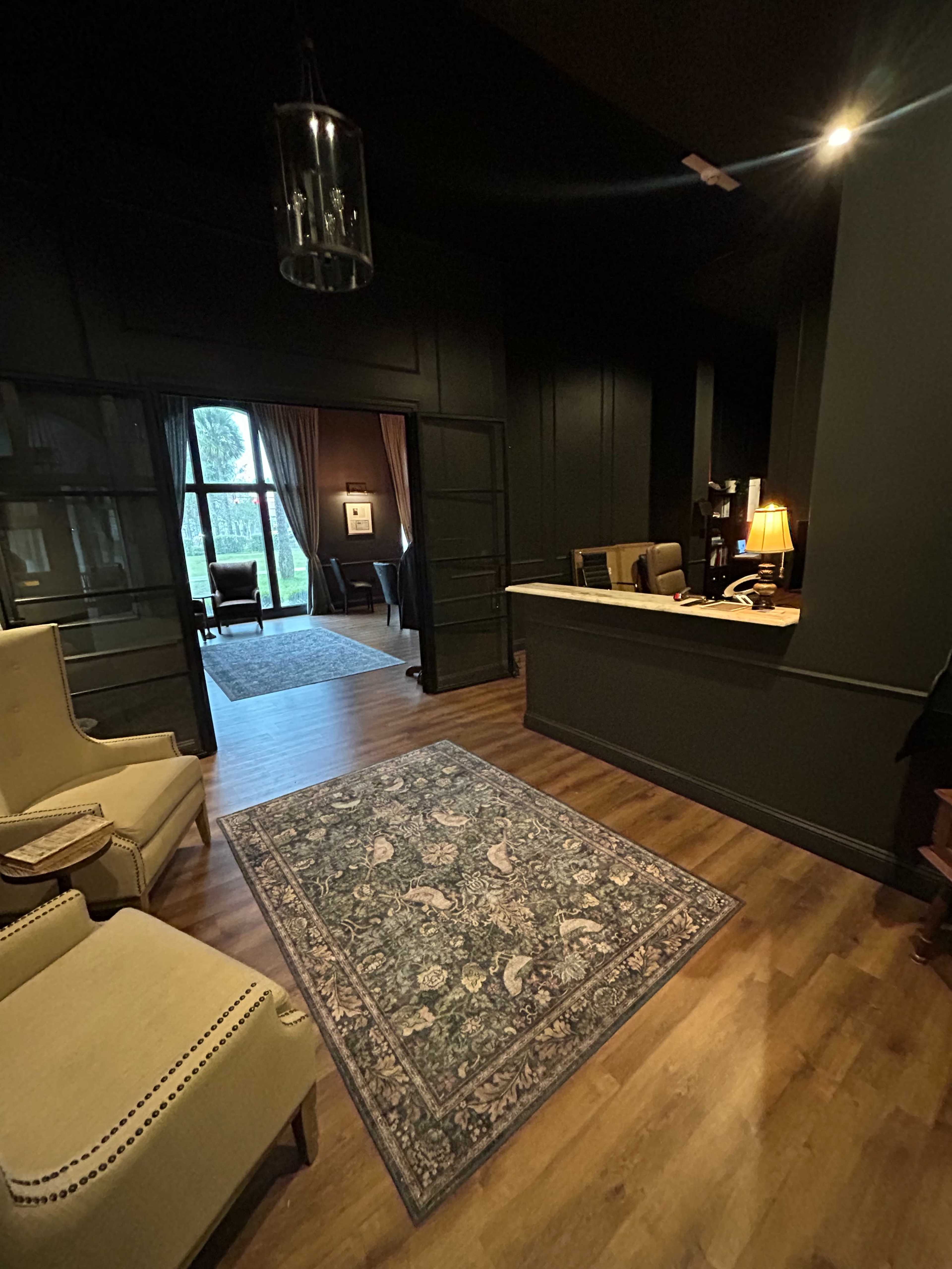 The image shows a dimly lit reception area featuring a rug, two armchairs, and a desk, with an open doorway leading to another room with a view of greenery outside.