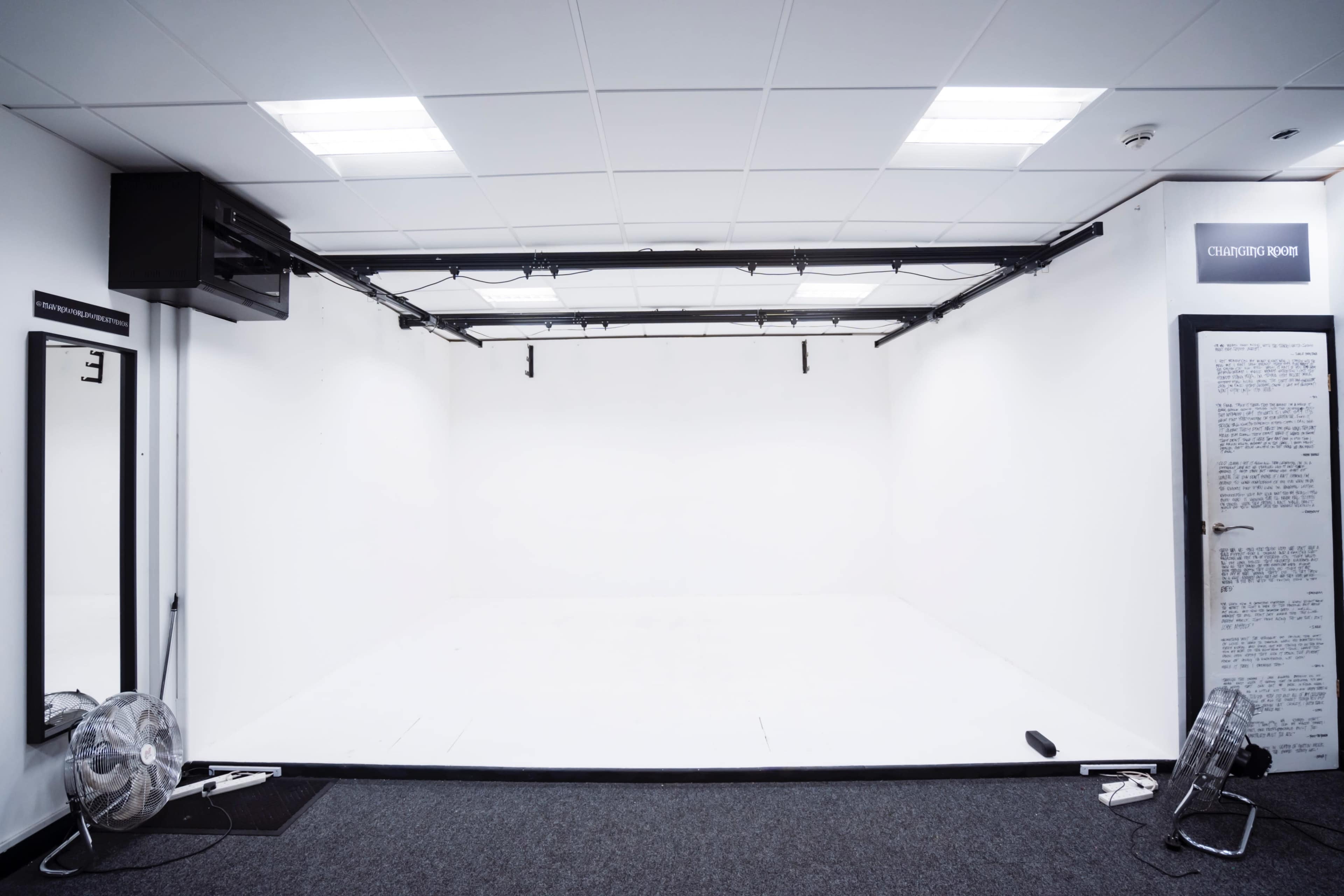 An empty studio space with white walls, a gray carpet, and a large mirror on the left.