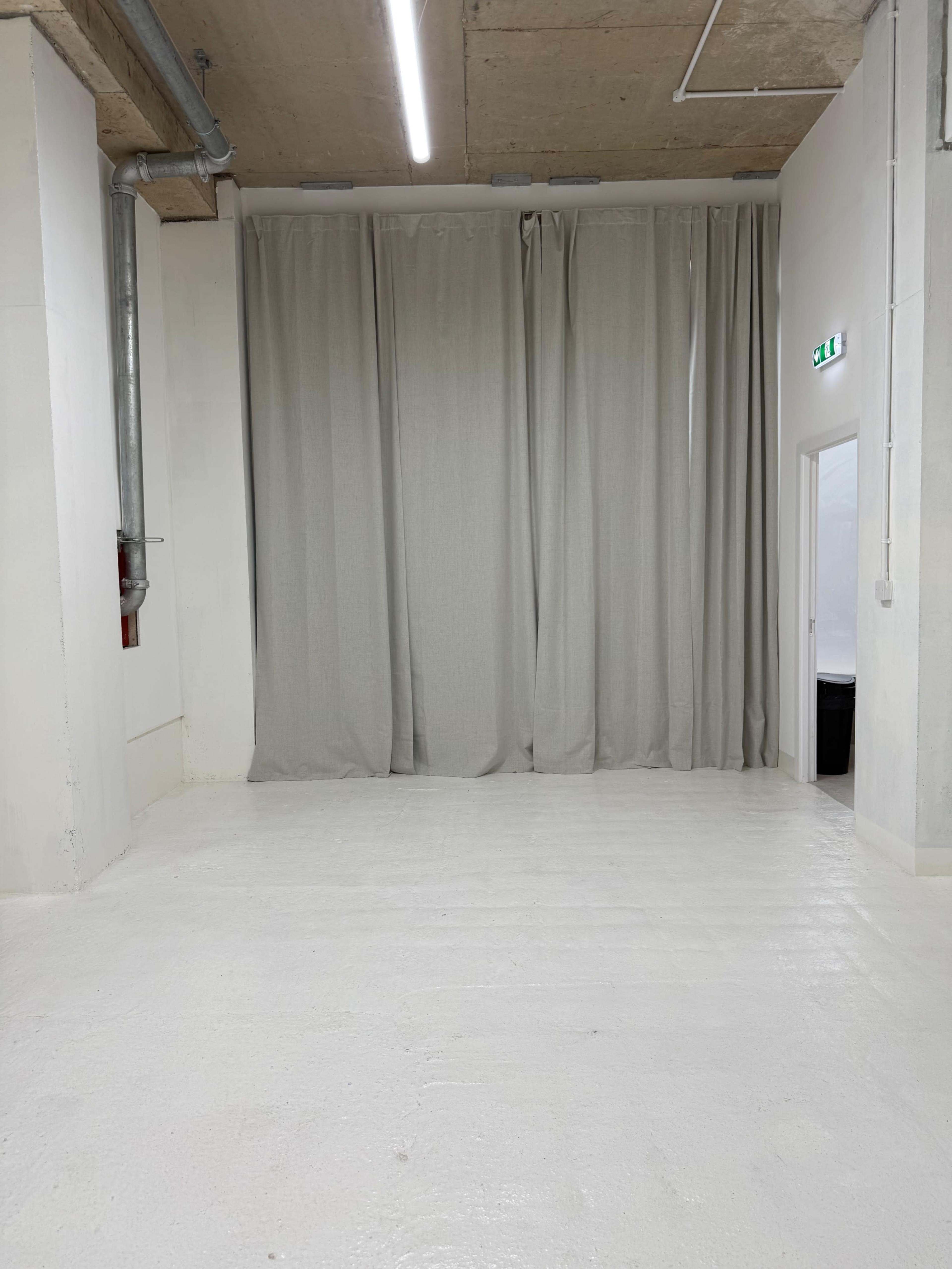 The image shows a spacious, minimalist room with white walls and flooring, featuring a large set of light-colored curtains along one side.
