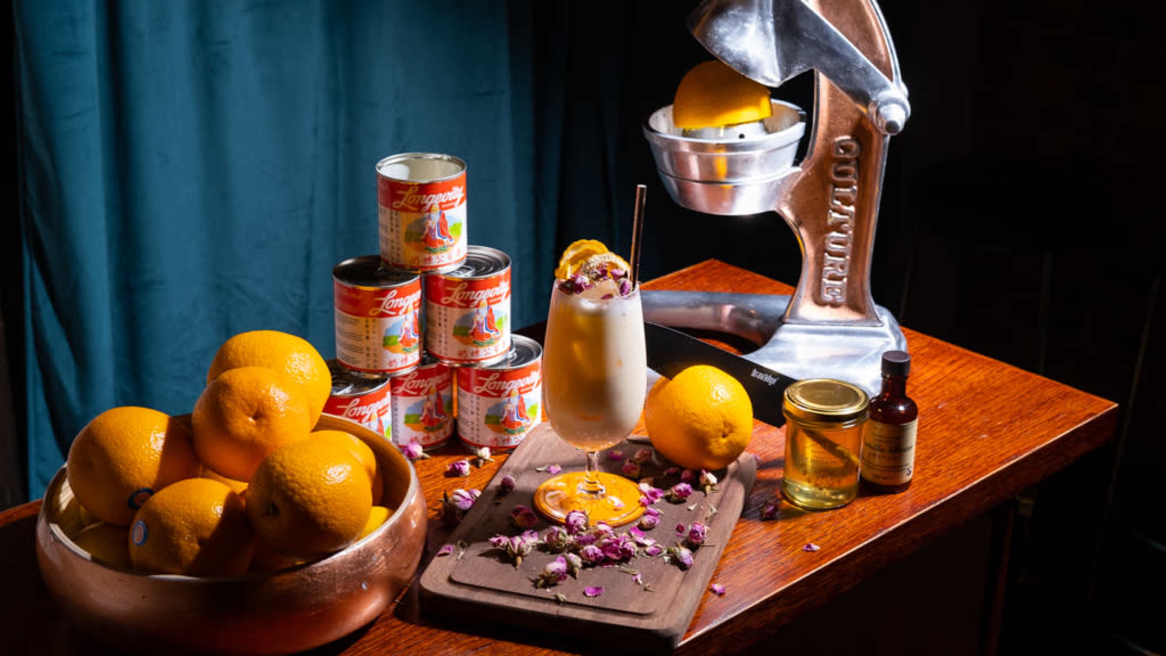The image features a neatly arranged display of oranges, cans of fruit, a cocktail with floral garnish, a wooden cutting board, and a metal citrus juicer.