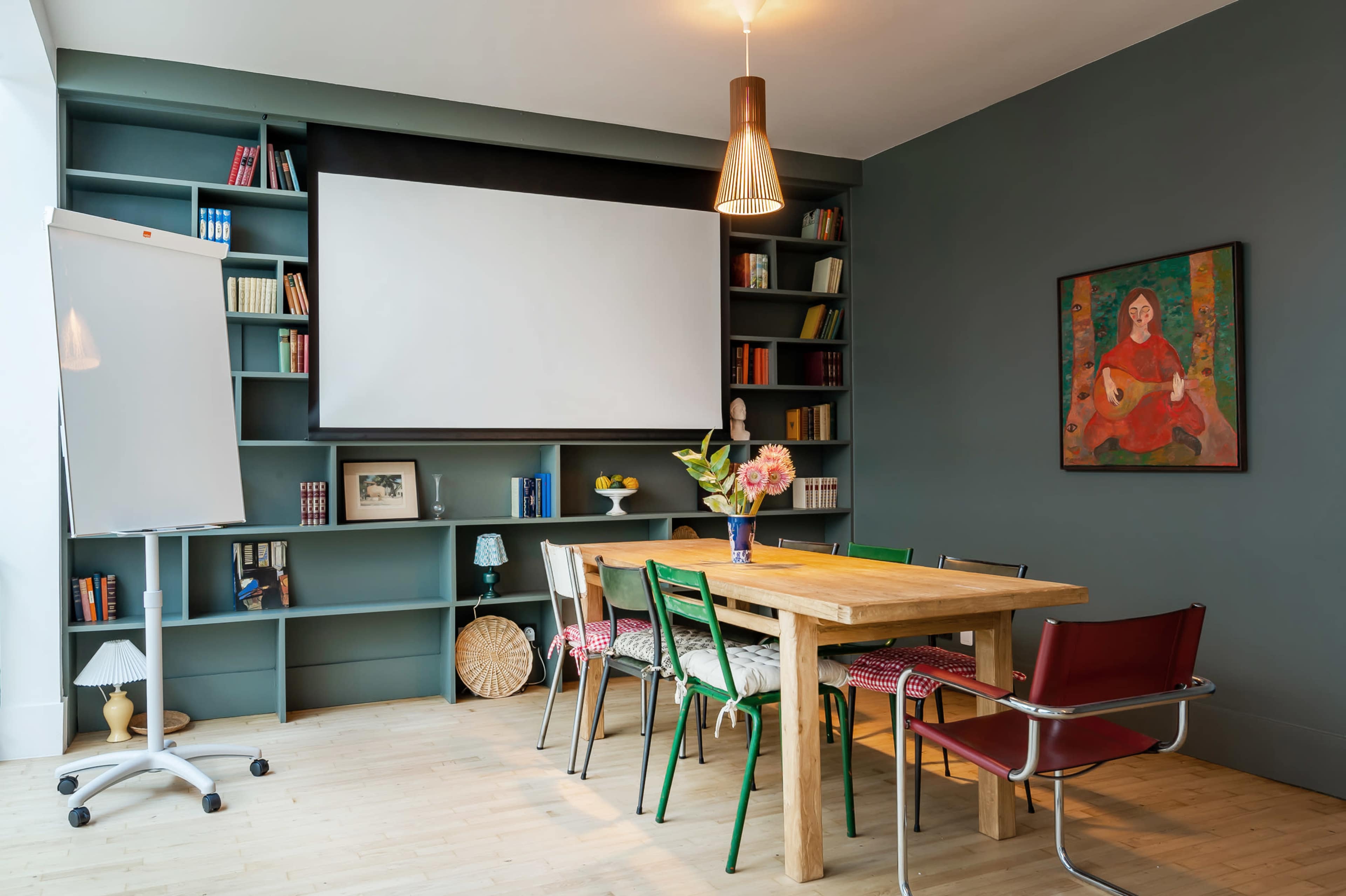 A modern meeting room features a long wooden table surrounded by colorful chairs, with a large projection screen and bookshelves filled with books in the background.