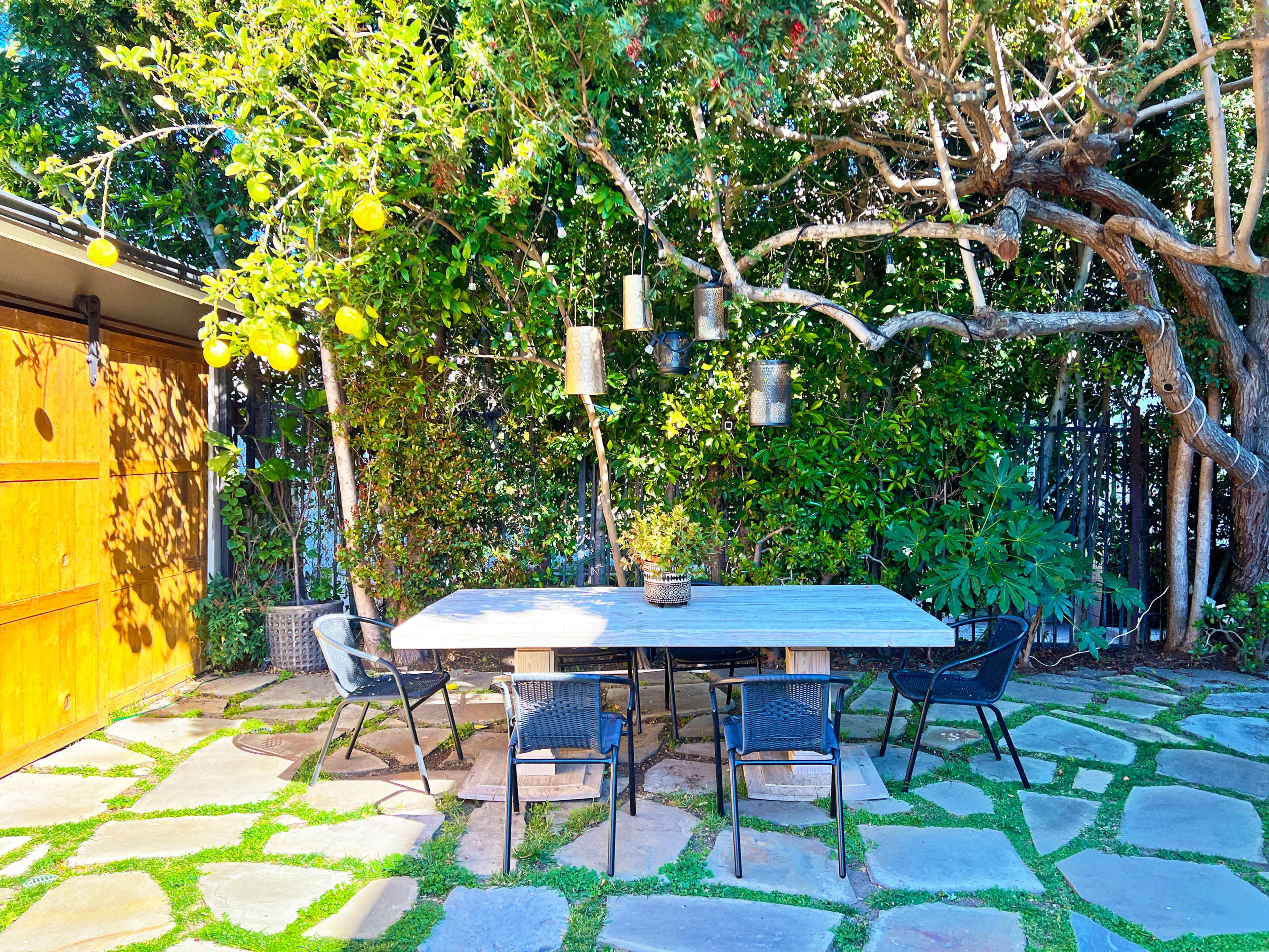 A wooden dining table with chairs is positioned on a stone patio beneath a tree with hanging lanterns and citrus fruits.