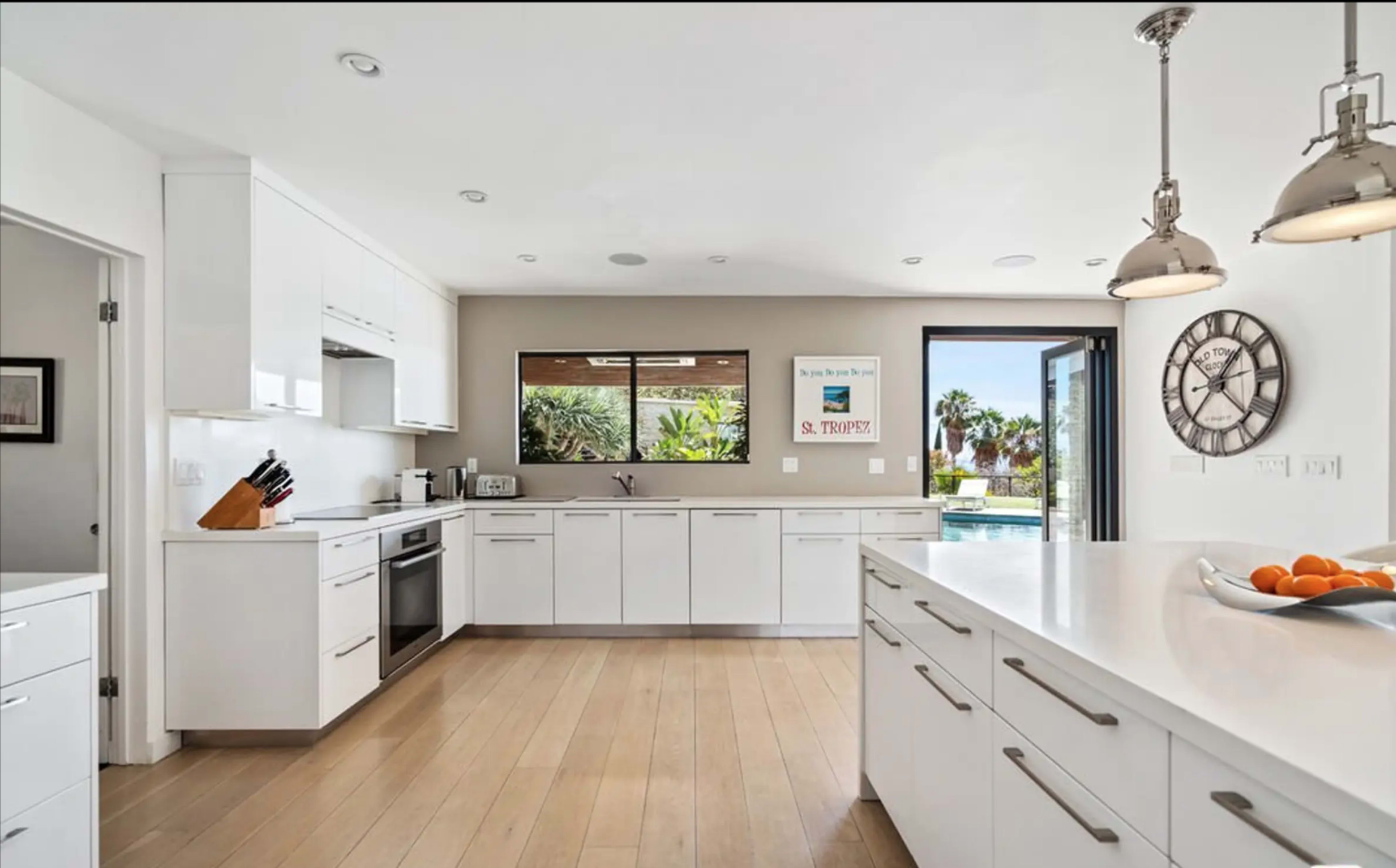 The image shows a modern kitchen with white cabinetry, a large island, and a view of an outdoor area through a large window.