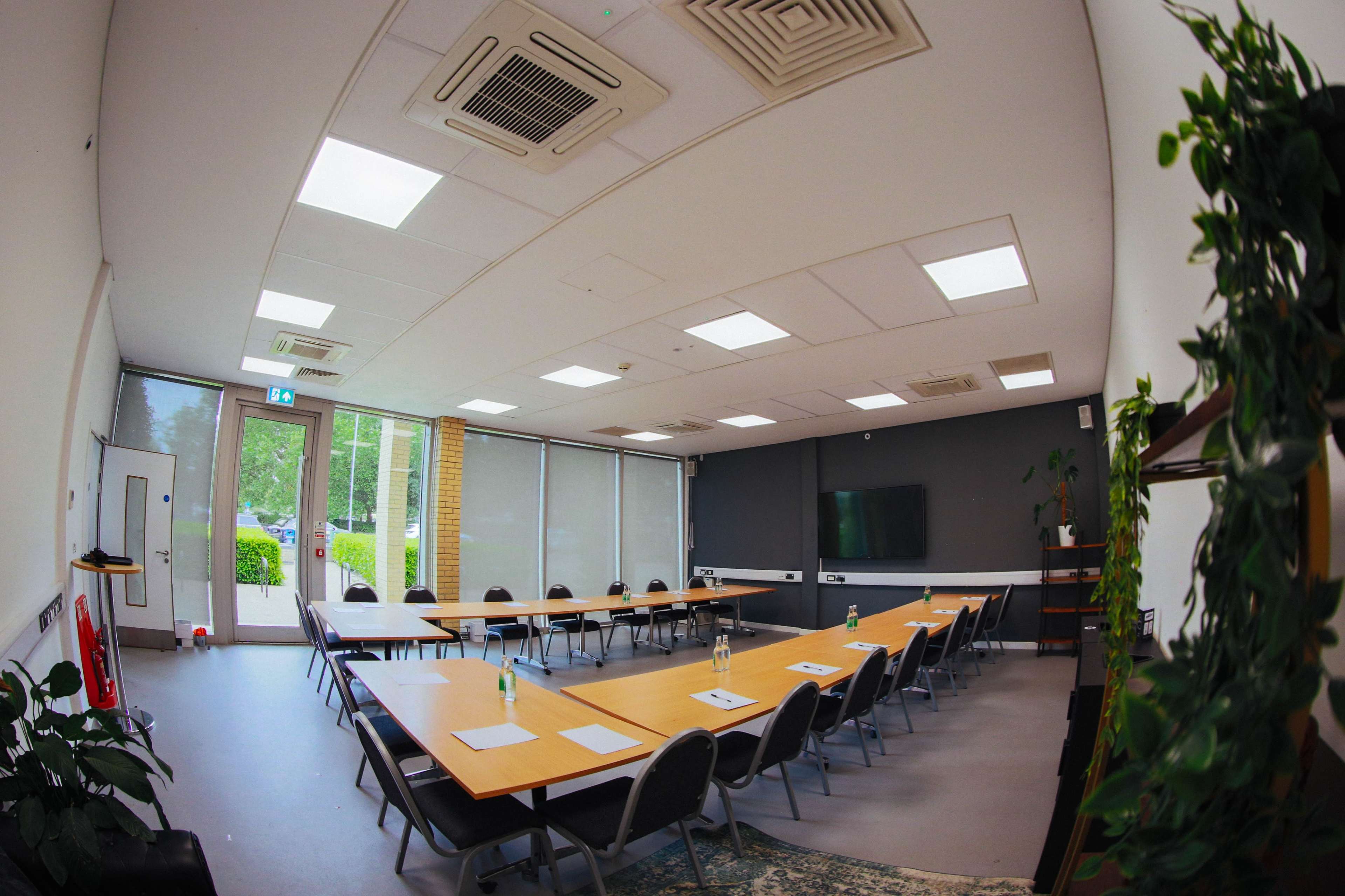 The image shows a modern conference room with a long table set up in a U-shape, surrounded by chairs, and featuring large windows that let in natural light.