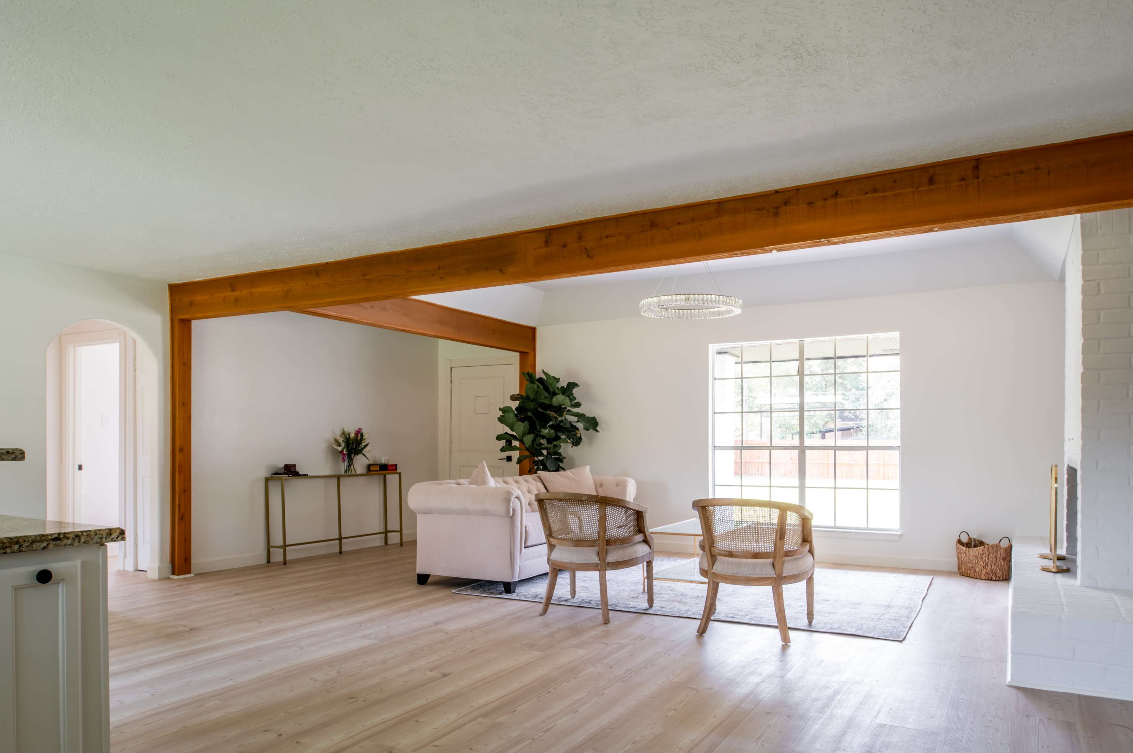A bright living room features a beige sofa, two wooden chairs, and a large window, with exposed wooden beams on the ceiling.